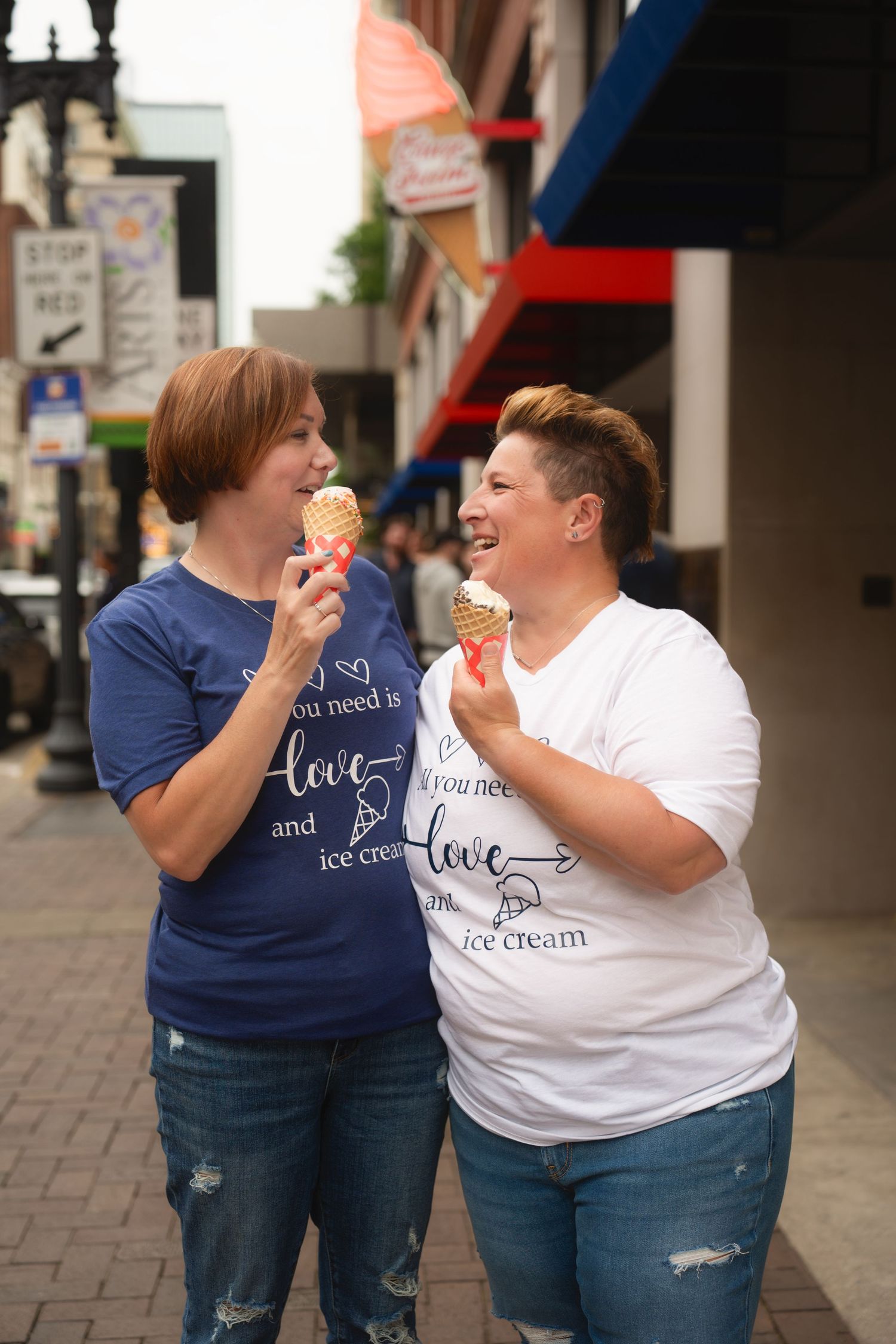 Two people in matching shirts share ice cream cones while laughing together on a city sidewalk.