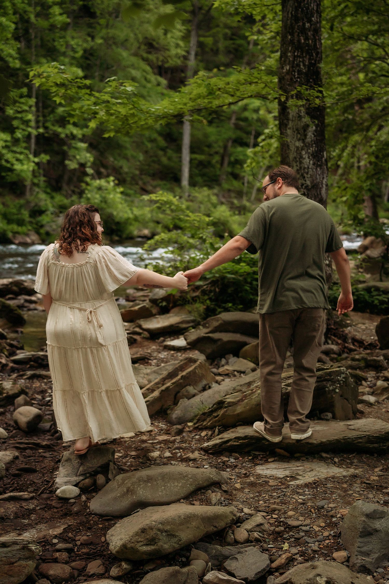 A couple walks carefully across rocky terrain near a stream in a lush forest setting.