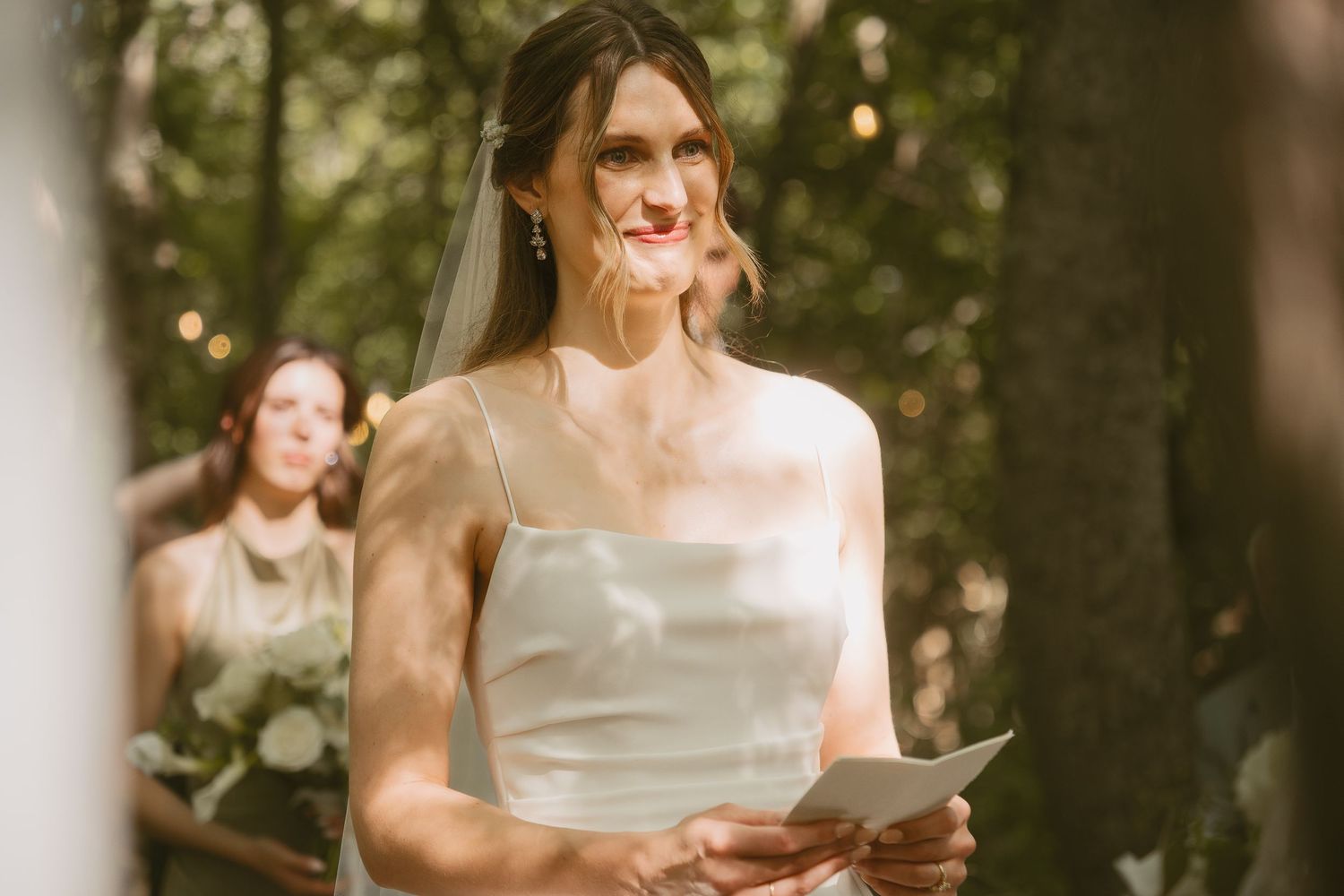 A person in a white dress reads from a paper during an outdoor ceremony in a wooded setting.