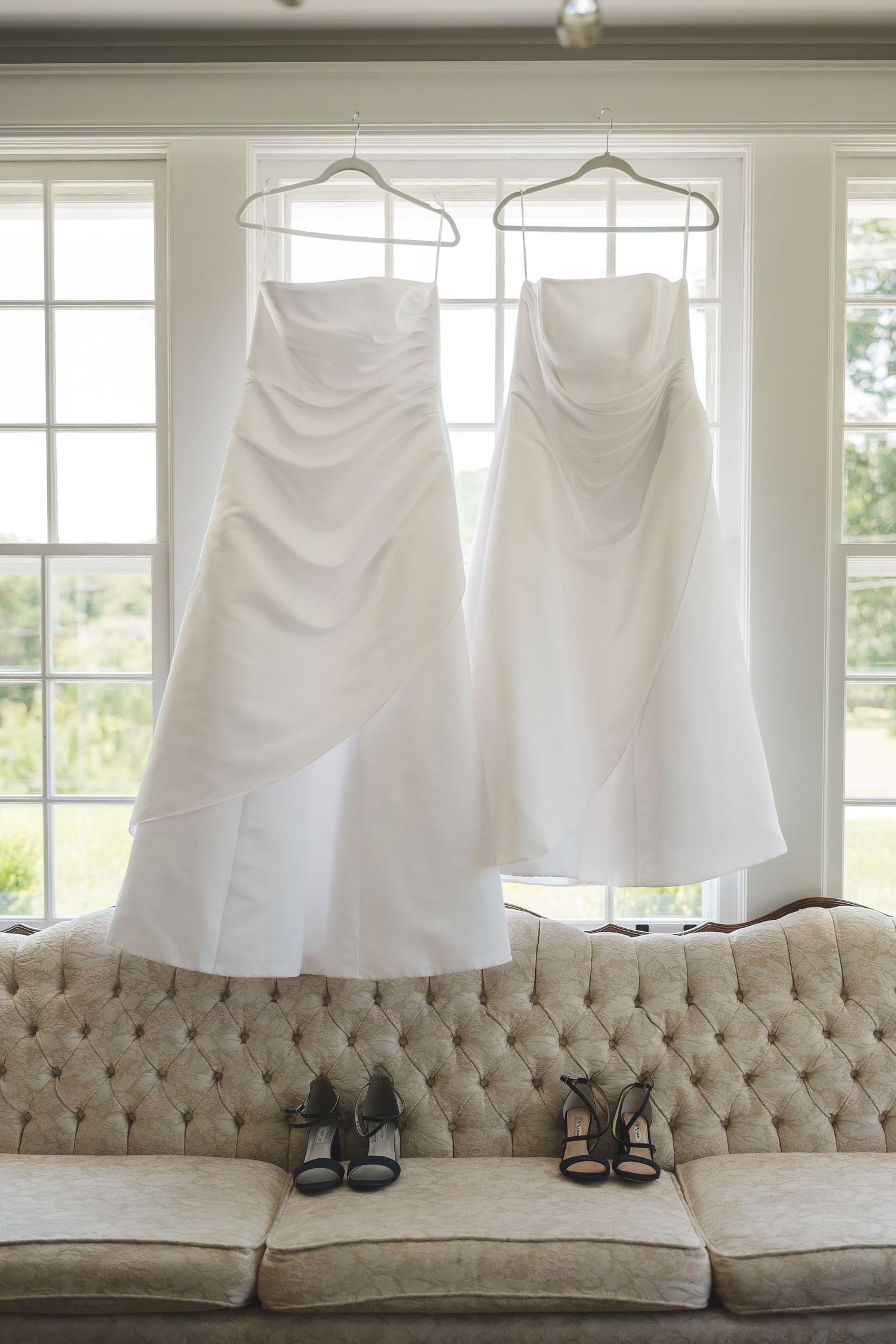 White wedding dresses hang above an elegant tufted beige couch with pairs of matching shoes arranged below.