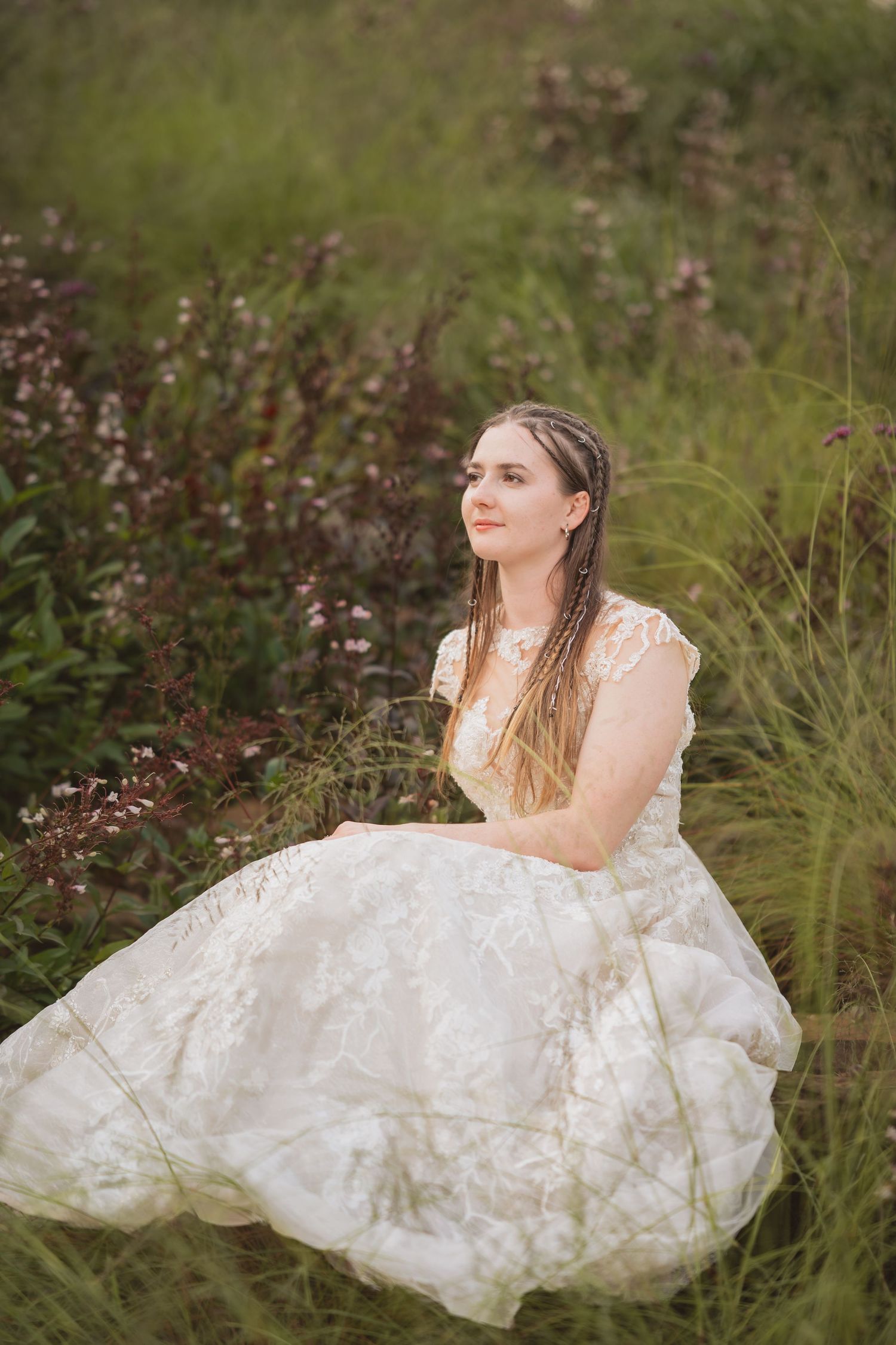 A bride in a white lace wedding gown reclines in a scenic meadow with wildflowers and tall grass.