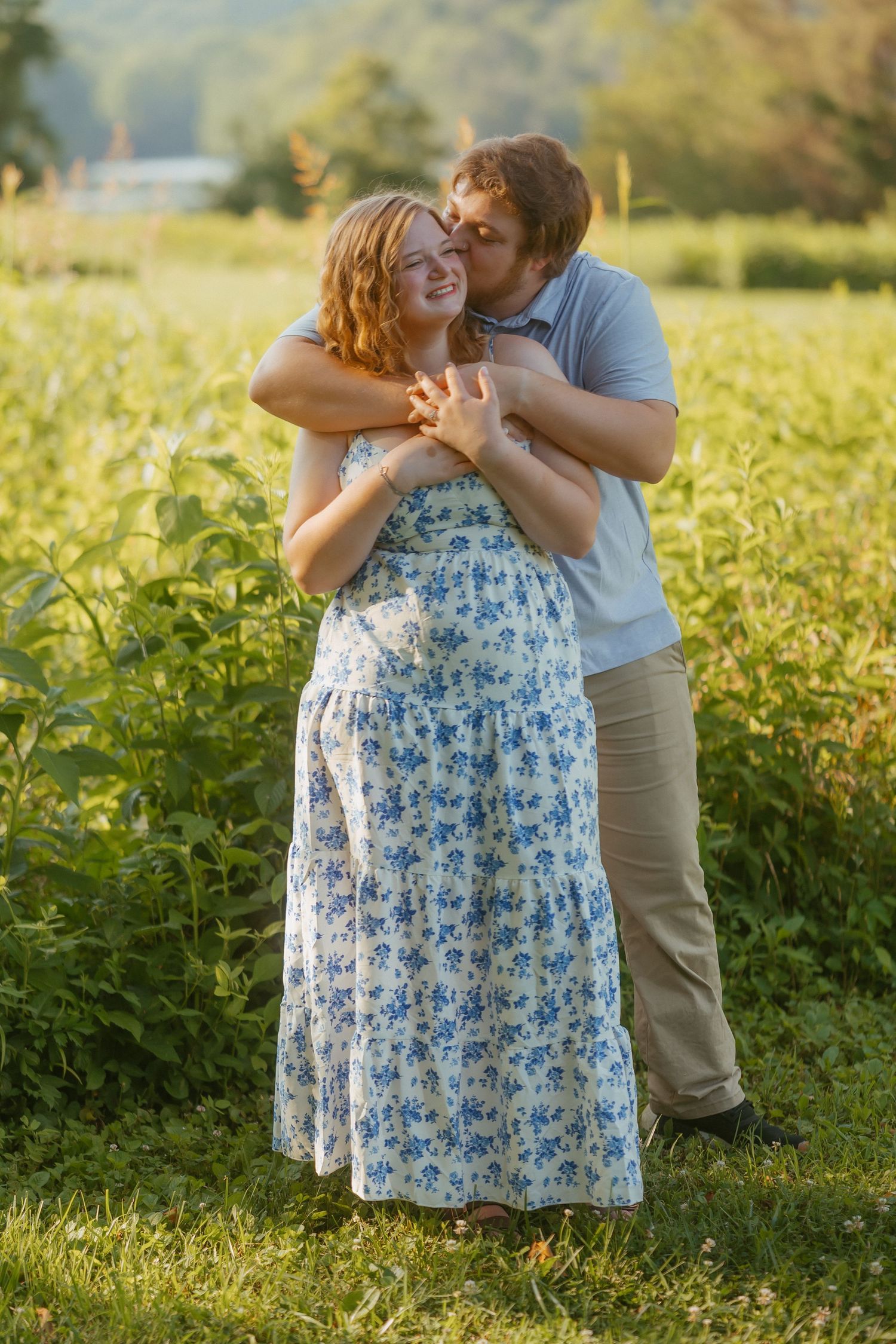 A couple embraces in a sunlit meadow wearing blue floral dress and gray clothing during a romantic outdoor photoshoot.
