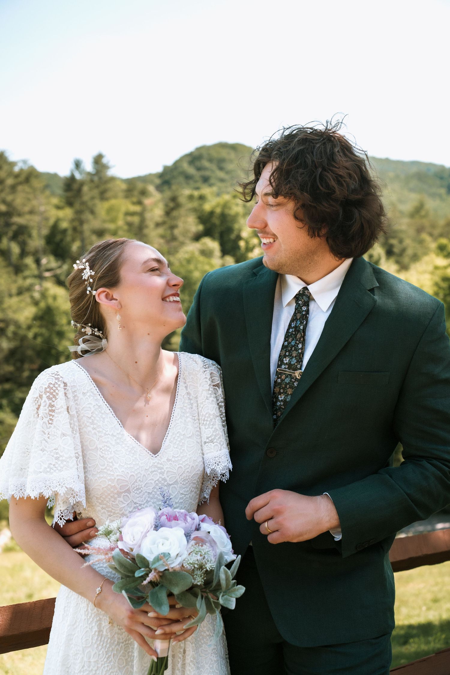 Newlywed couple shares joyful laughter at outdoor wedding ceremony with mountains and trees in background.