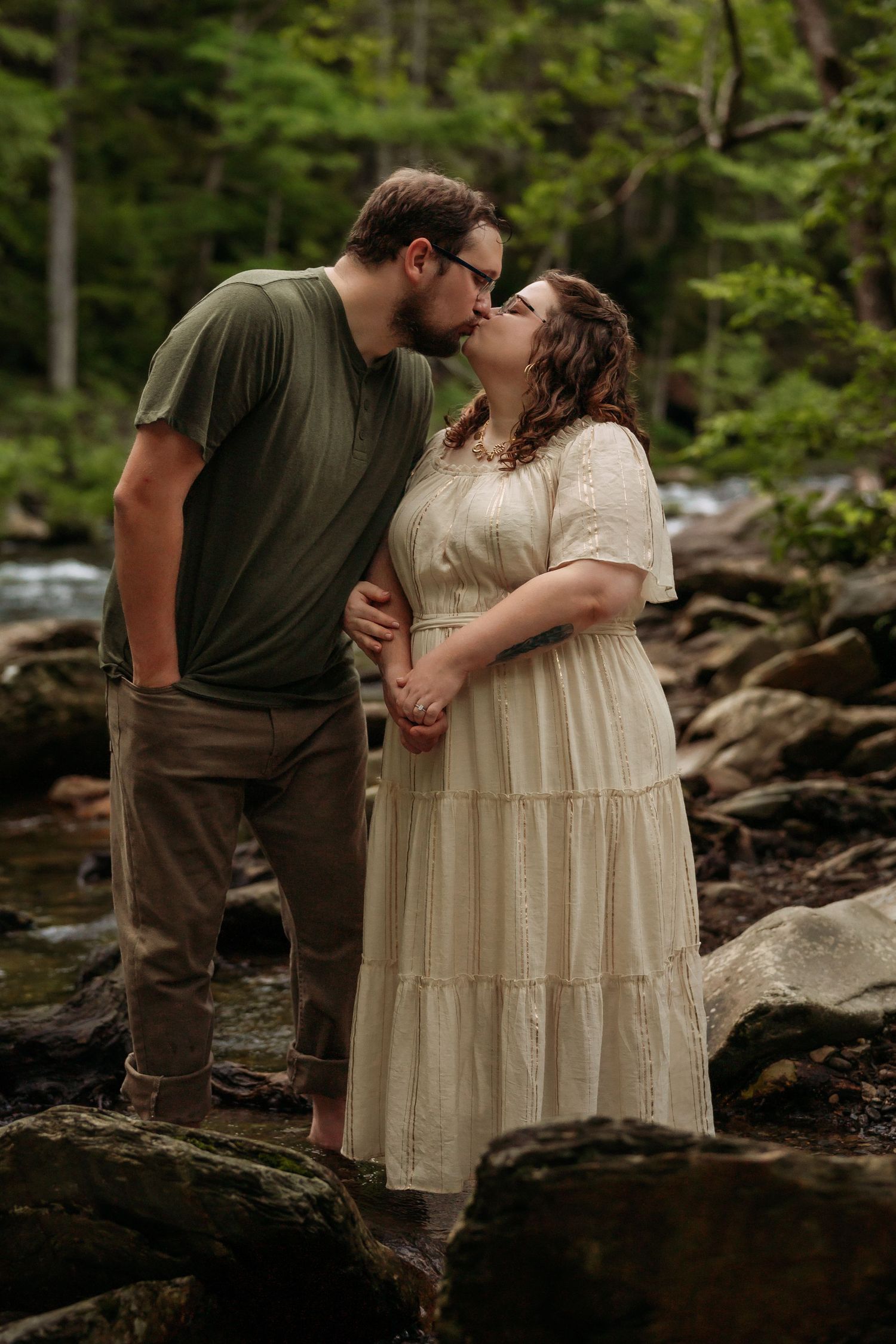Couple embracing on rocky riverbank surrounded by forest landscape.