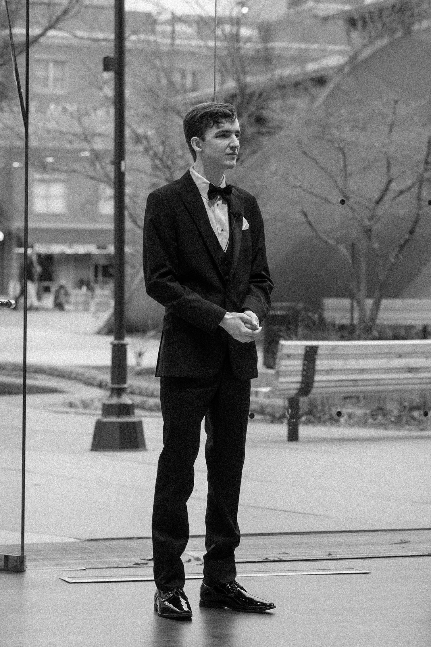 A vintage black and white photograph of someone in formal attire with a bow tie standing outdoors in an urban setting.