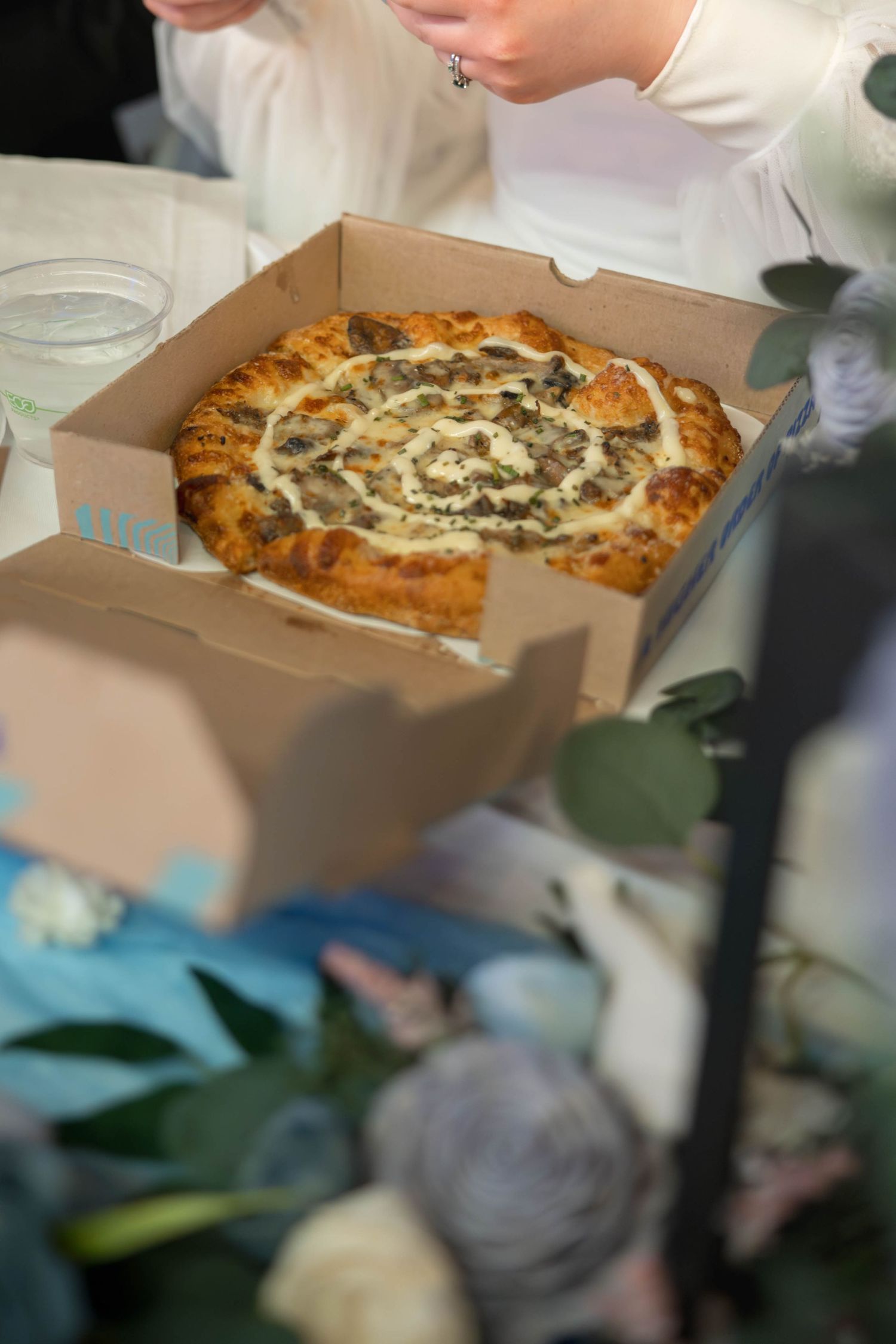 A gourmet pizza with white sauce in a takeout box surrounded by wedding decorations.