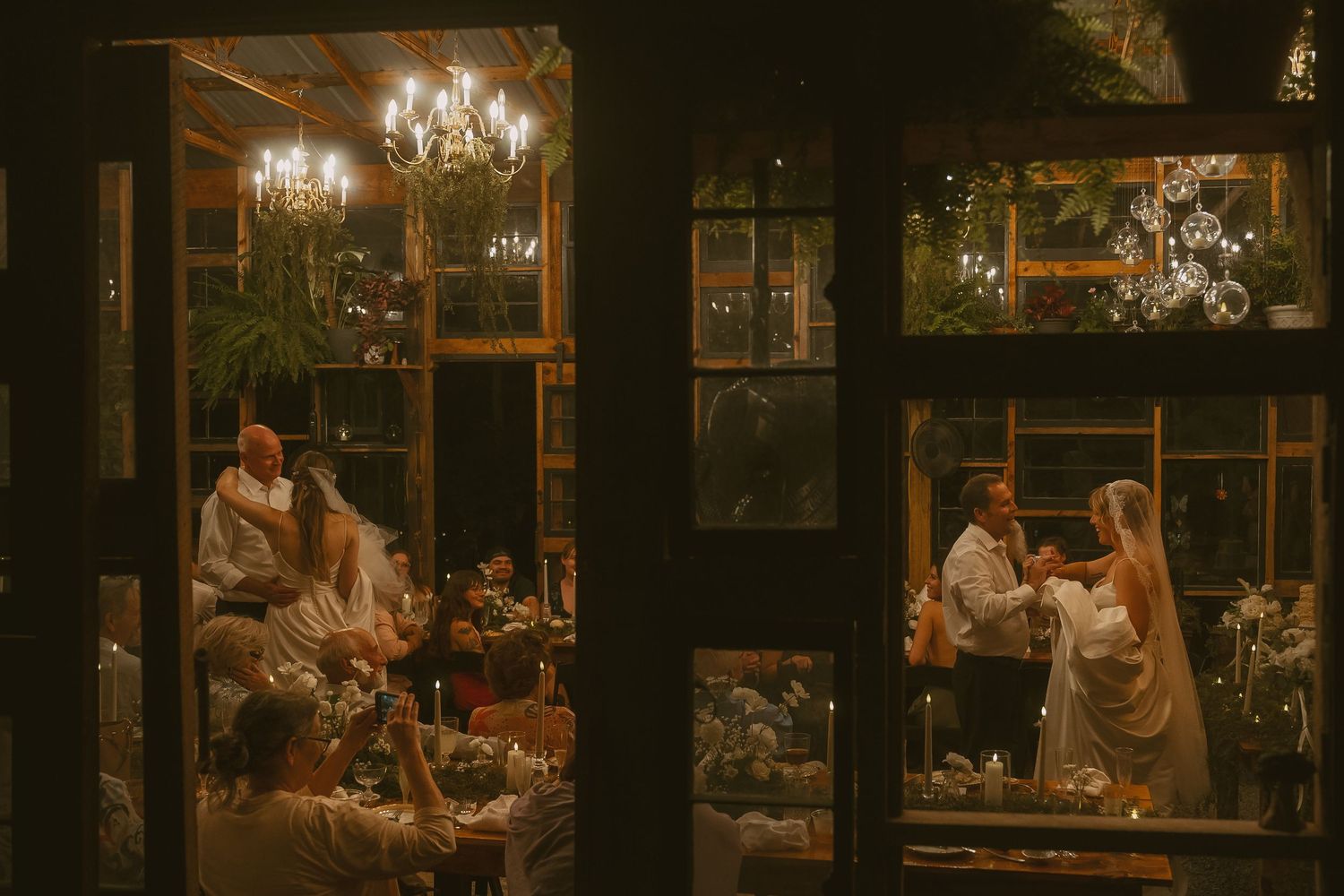 Warm glowing chandeliers illuminate a rustic indoor reception venue with dark wooden beams.