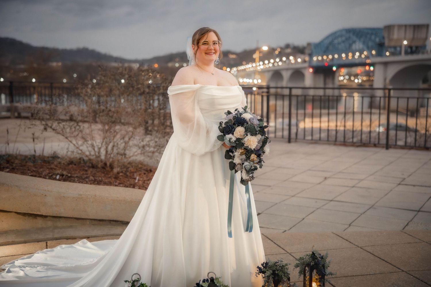 A bride in a flowing off-shoulder wedding gown poses with a blue and white bouquet on an outdoor terrace at dusk.