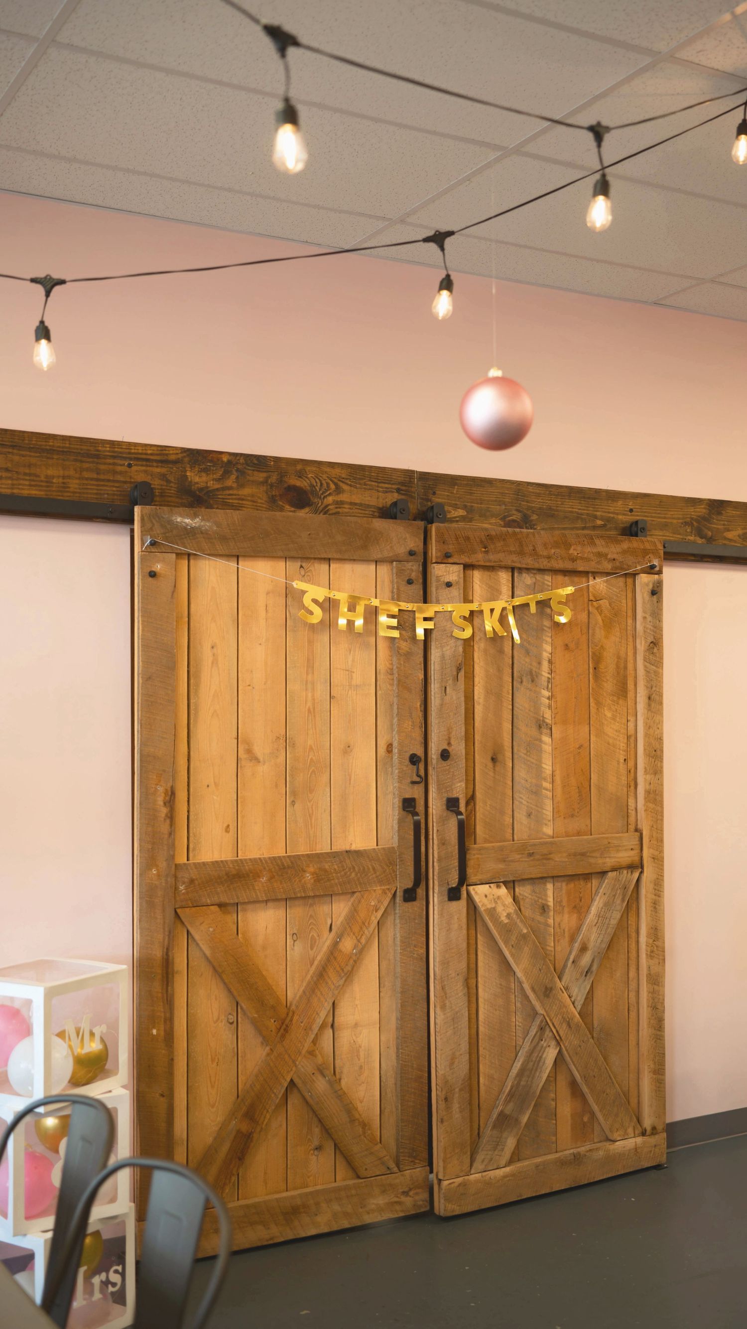 Rustic wooden barn doors with sliding hardware and string lights hang in an interior space with pink walls.