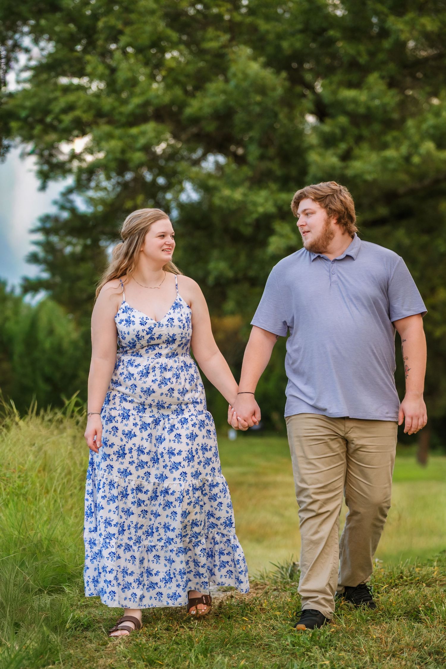 A couple holds hands while walking through a lush green field on a summer day, wearing casual attire.