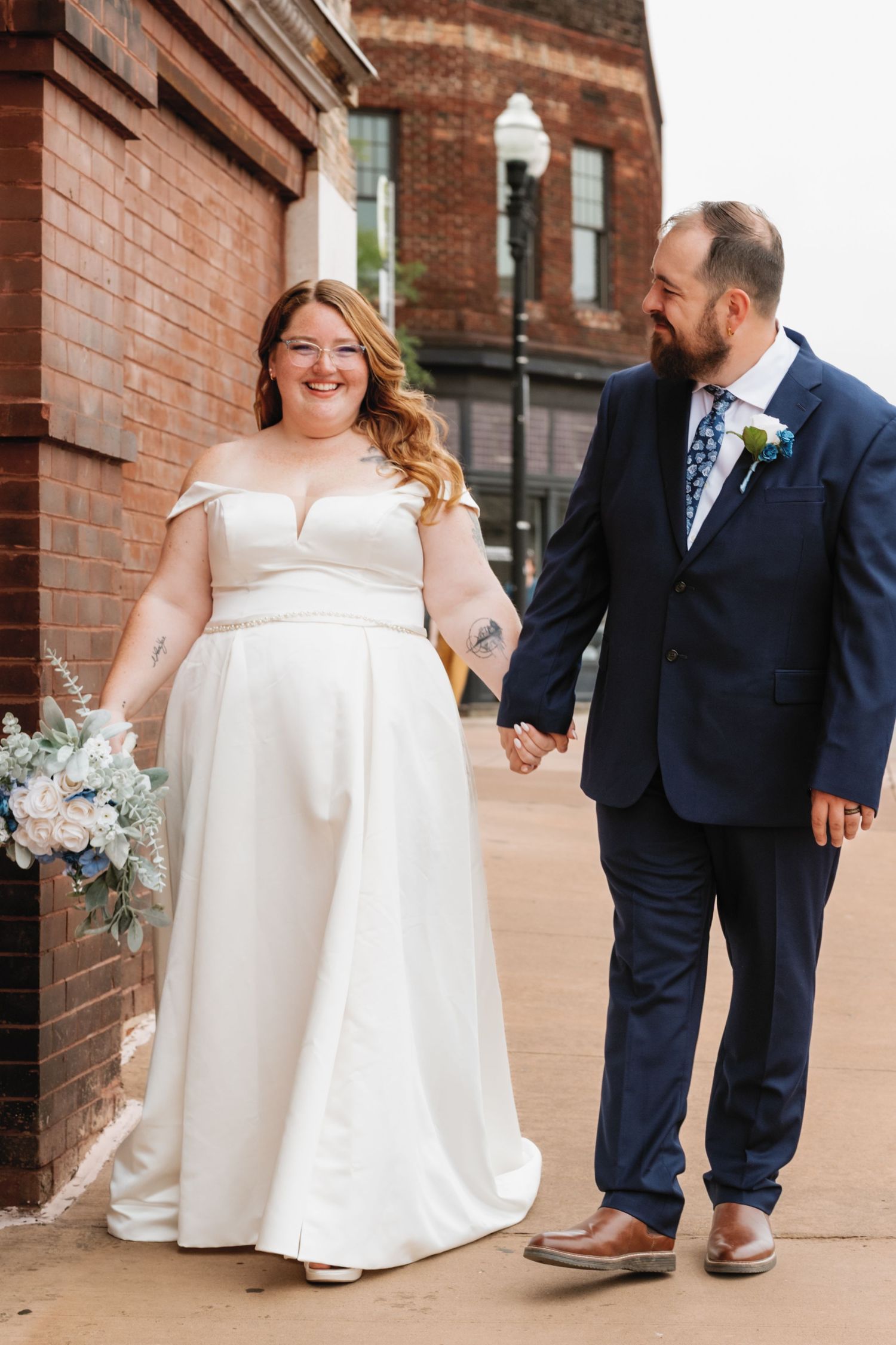Couple on their wedding day holding hands and smiling while walking along a brick building exterior.