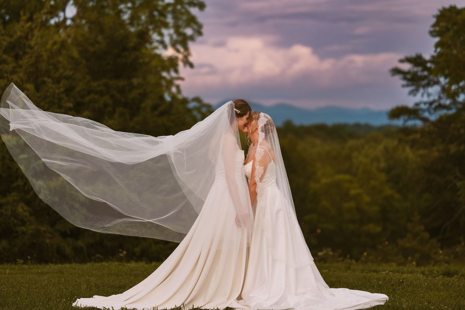 A dramatic wedding dress with flowing veil billows in the wind against a mountain landscape at sunset.