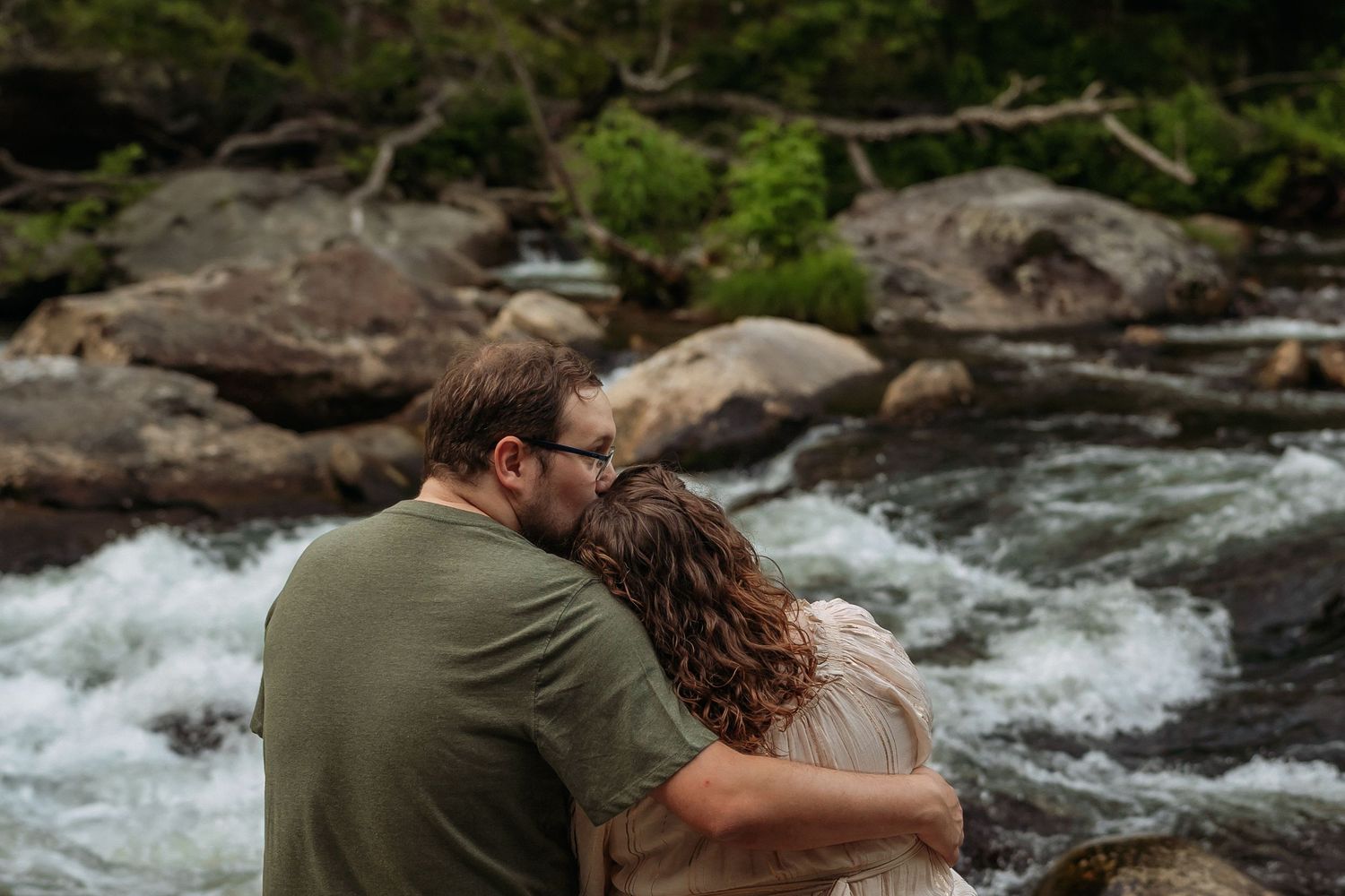 Two people embrace while standing near a rushing river surrounded by mossy rocks and flowing water.