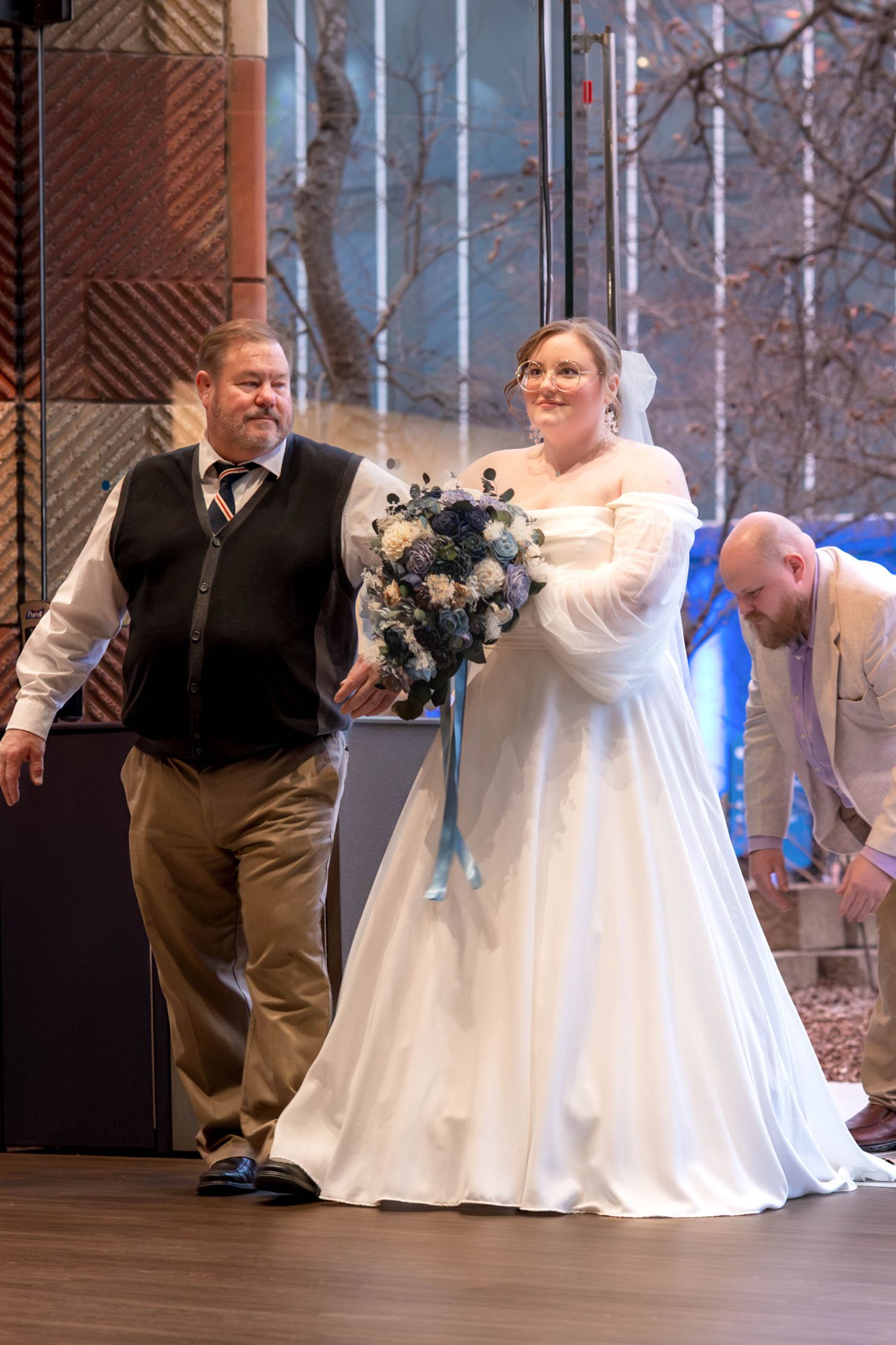 Bride in elegant white dress and fur wrap walks across stage holding blue and white bouquet during winter wedding ceremony.