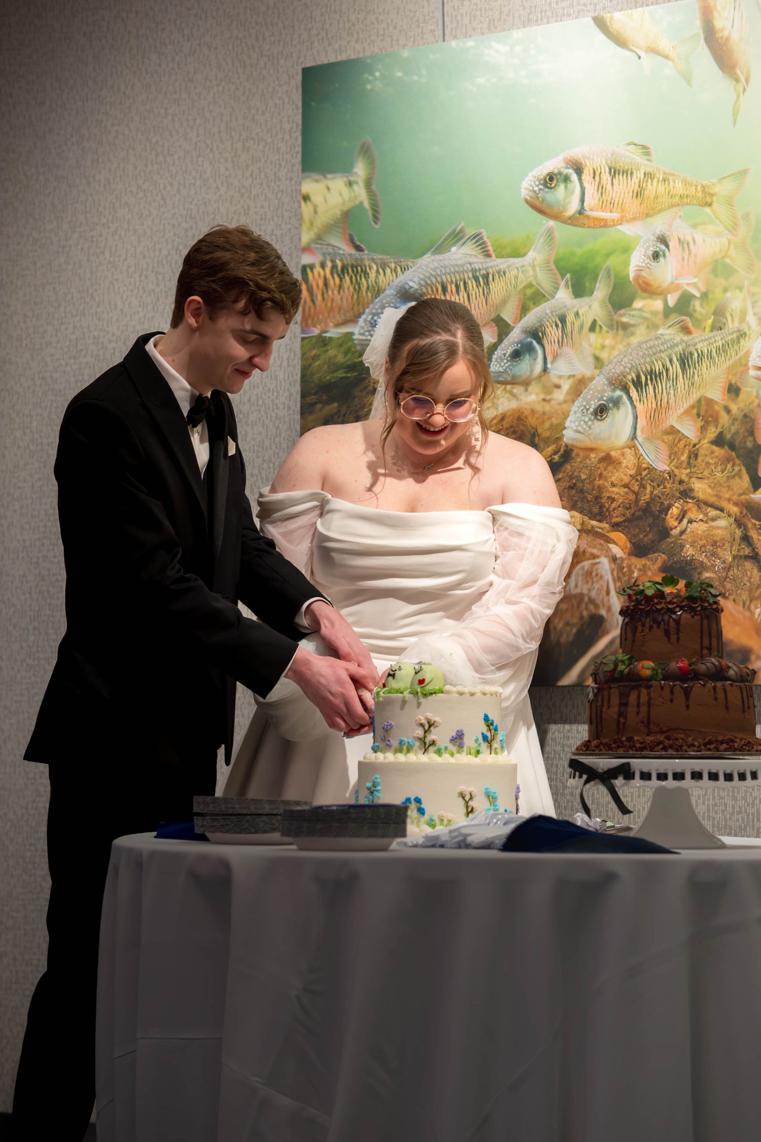 A couple in formal wedding attire cuts their white wedding cake together in front of a colorful aquatic-themed mural.