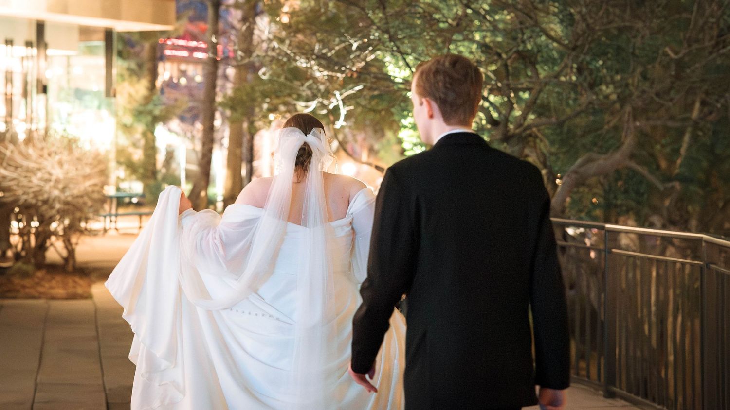 A couple in wedding attire walk together on a lit pathway during evening hours.