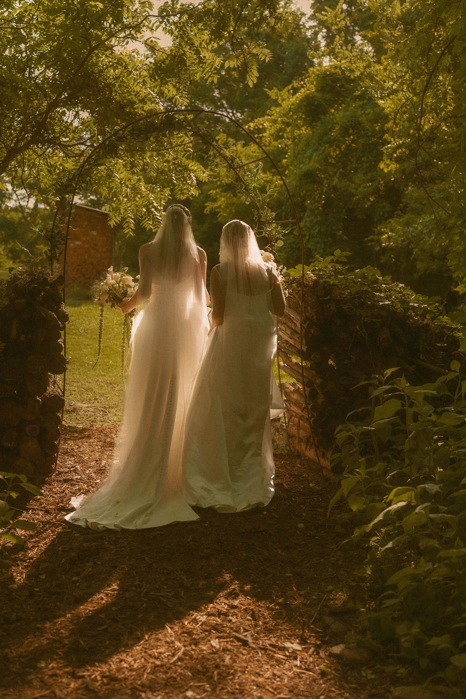 Two white wedding dresses casting ethereal silhouettes along a sunlit forest path.