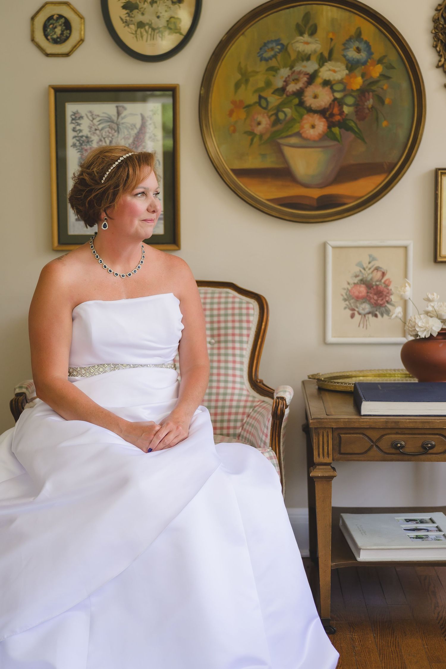 A woman in a white strapless wedding gown sits elegantly in an antique chair with floral artwork on the wall.