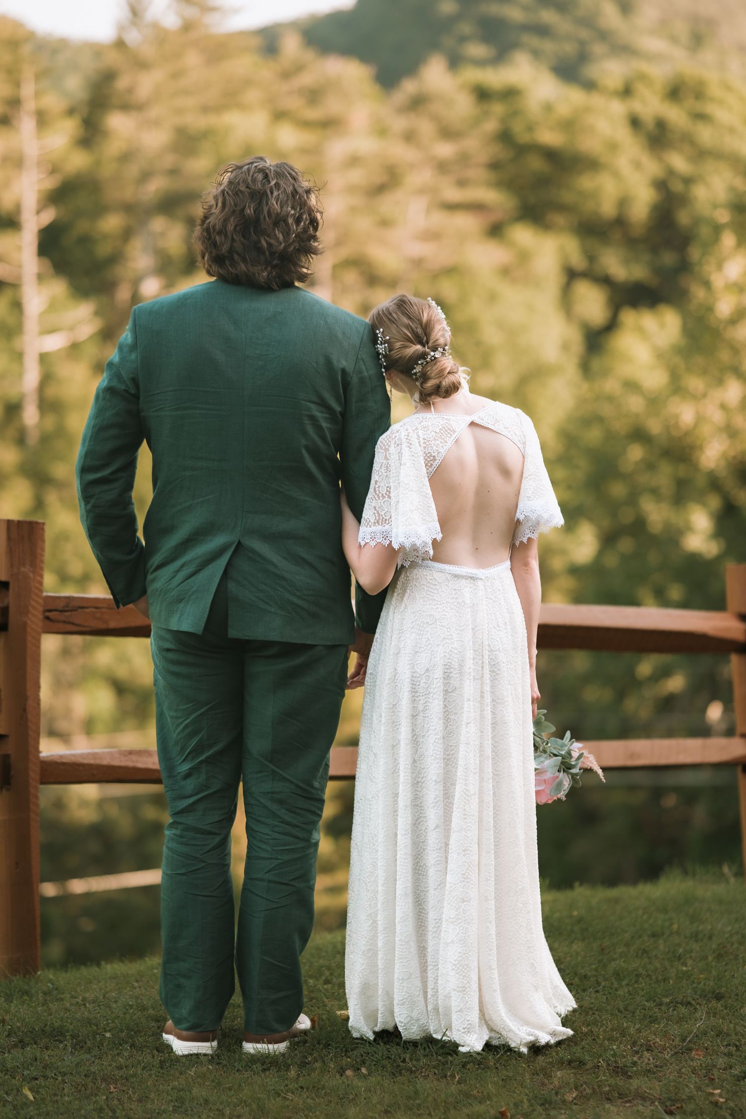 Two people in formal attire stand side by side on a grassy hill overlooking autumn trees at sunset.