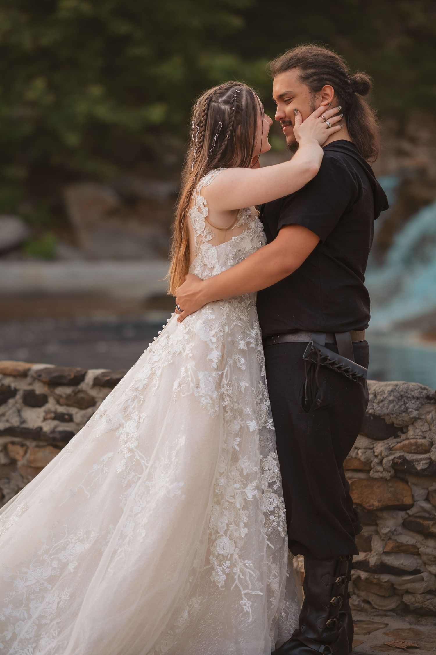 A couple in wedding attire embrace dramatically beside a stone wall during an outdoor photo session.