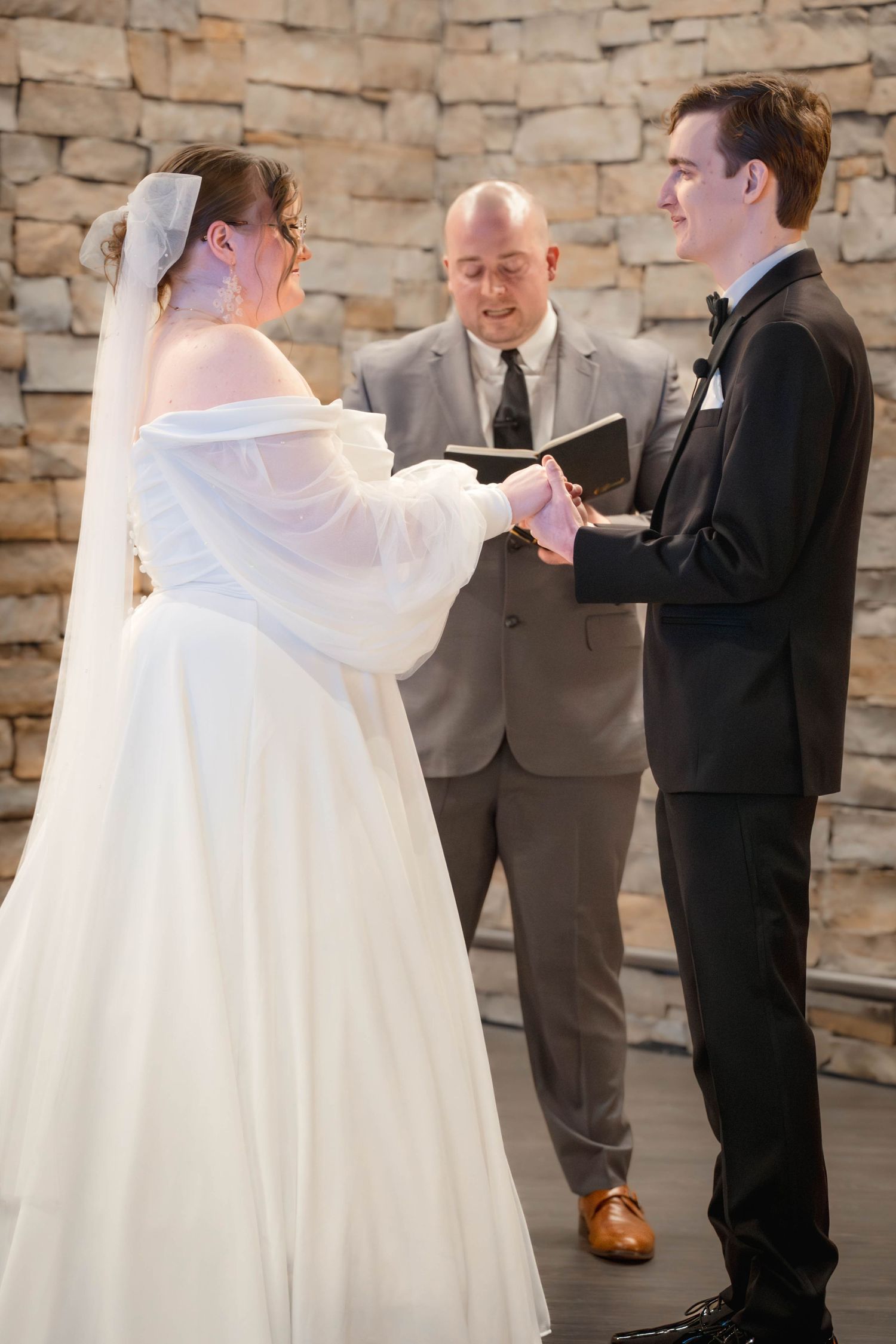 A wedding ceremony takes place with a bride in white gown and groom in black suit exchanging vows before an officiant.