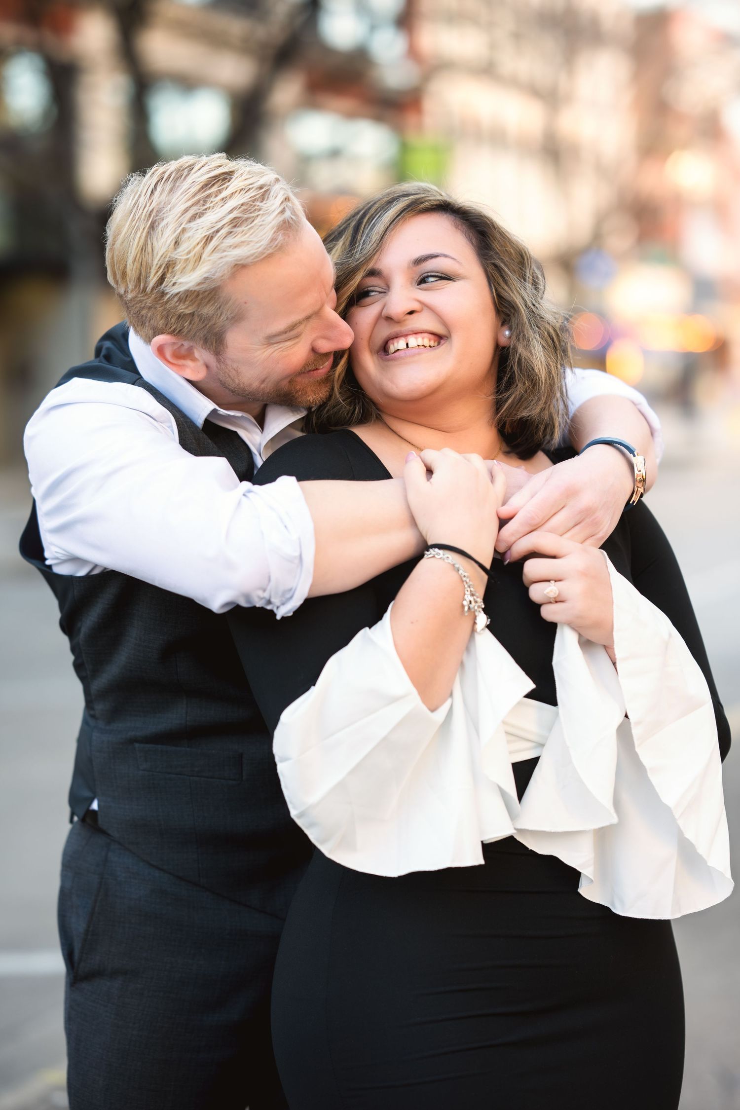 A joyful couple embraces and laughs together during a formal portrait session on a city street.