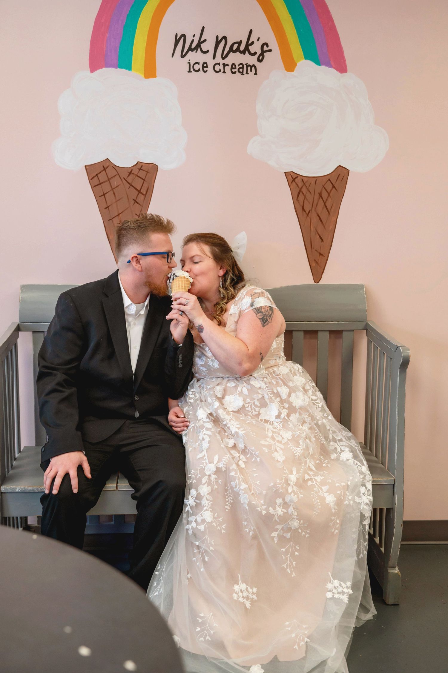A couple shares a tender moment on a bench beneath colorful rainbow and ice cream cone wall art at Nik Nak's Ice Cream.