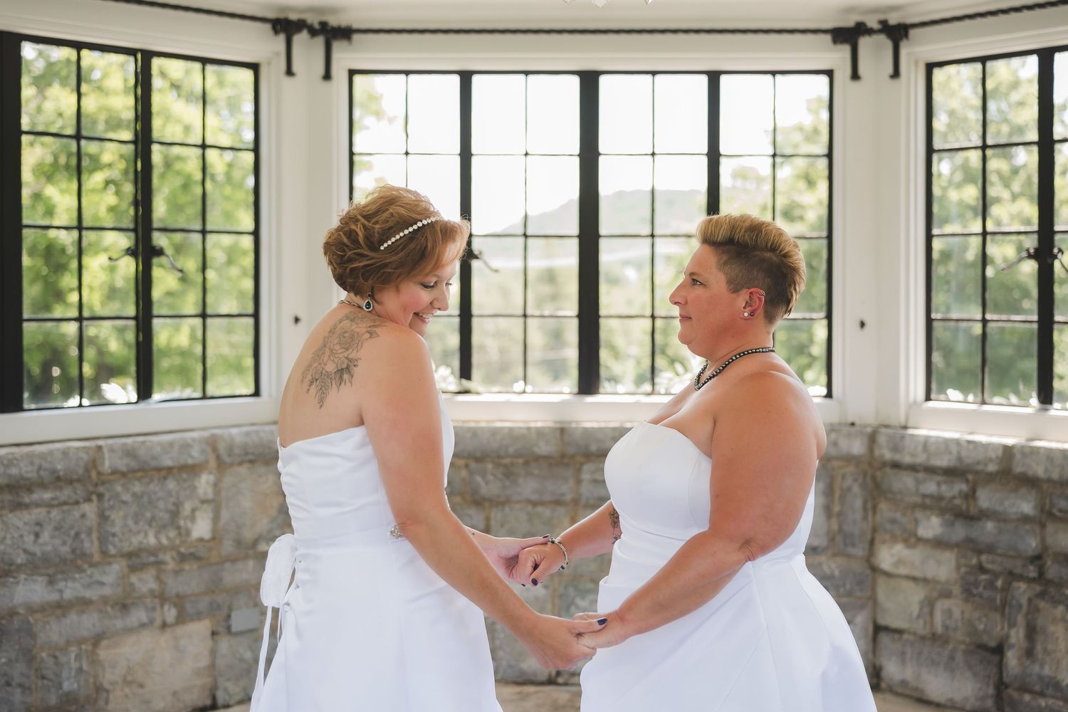 Two people in white wedding attire stand by bright windows in a stone-walled room during an intimate ceremony.