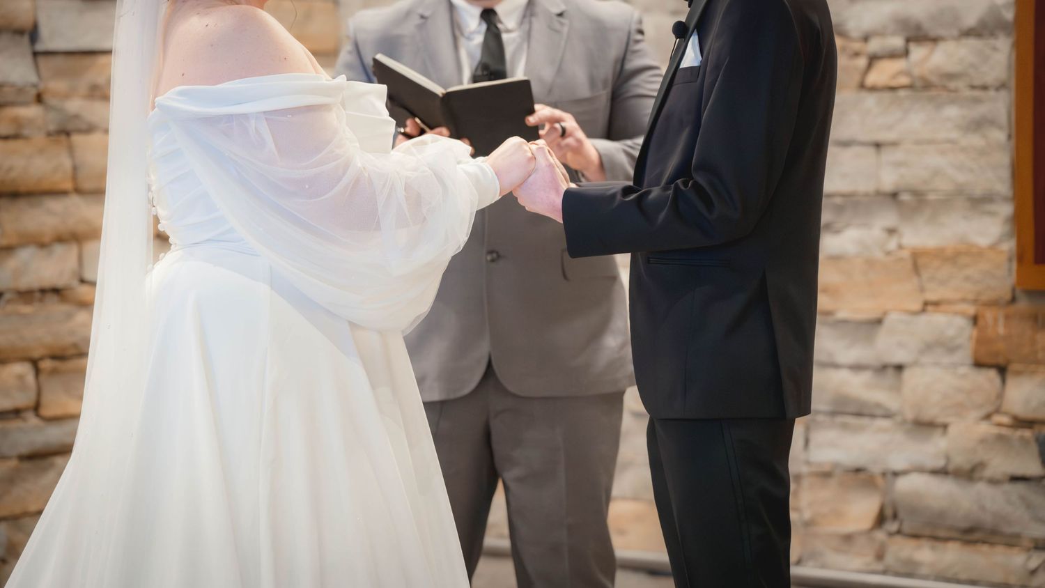 A couple holds hands during an intimate wedding ceremony against a rustic stone wall backdrop in matching formal attire.