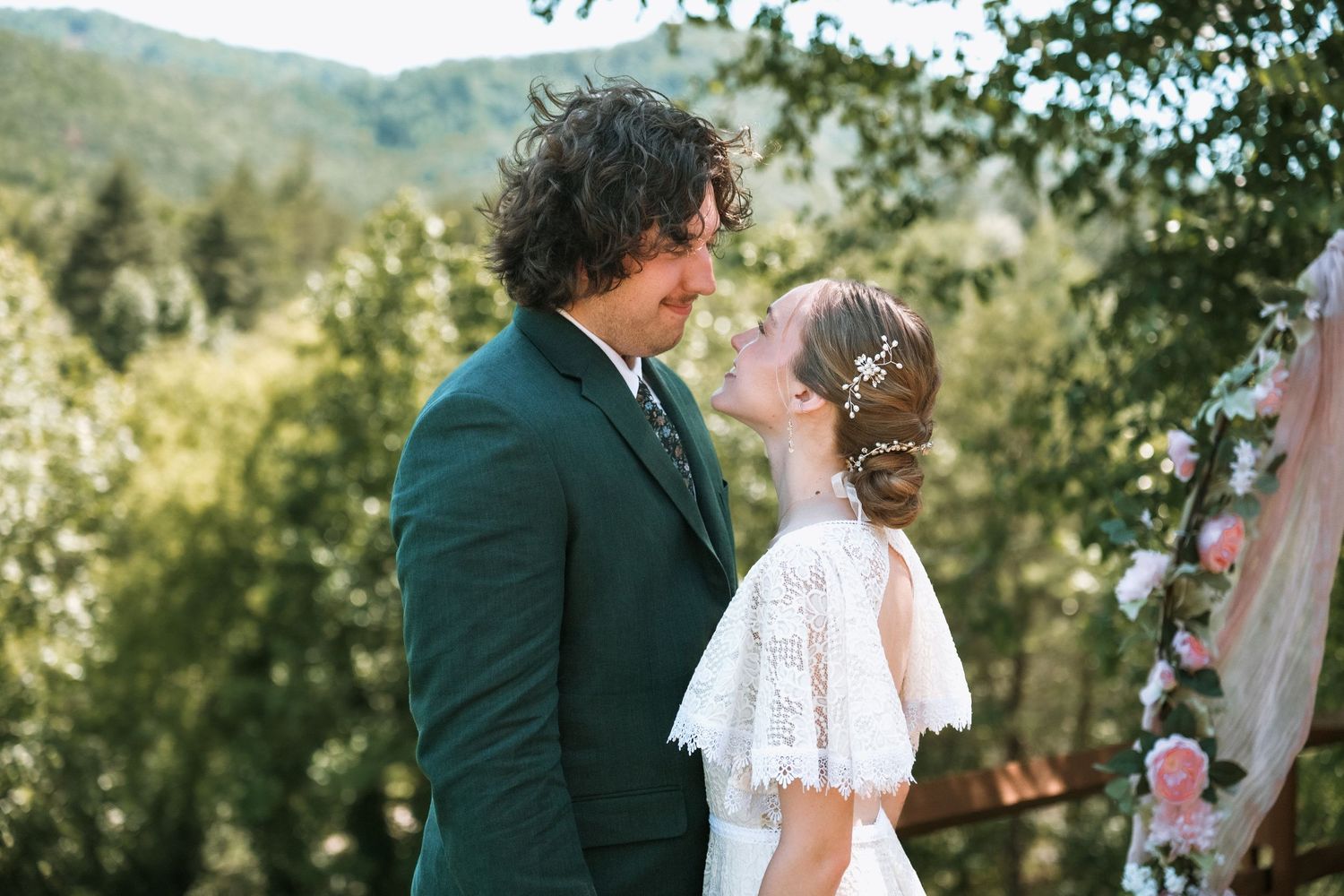 A happy couple shares a tender moment at an outdoor wedding ceremony with mountain and forest backdrop scenery.