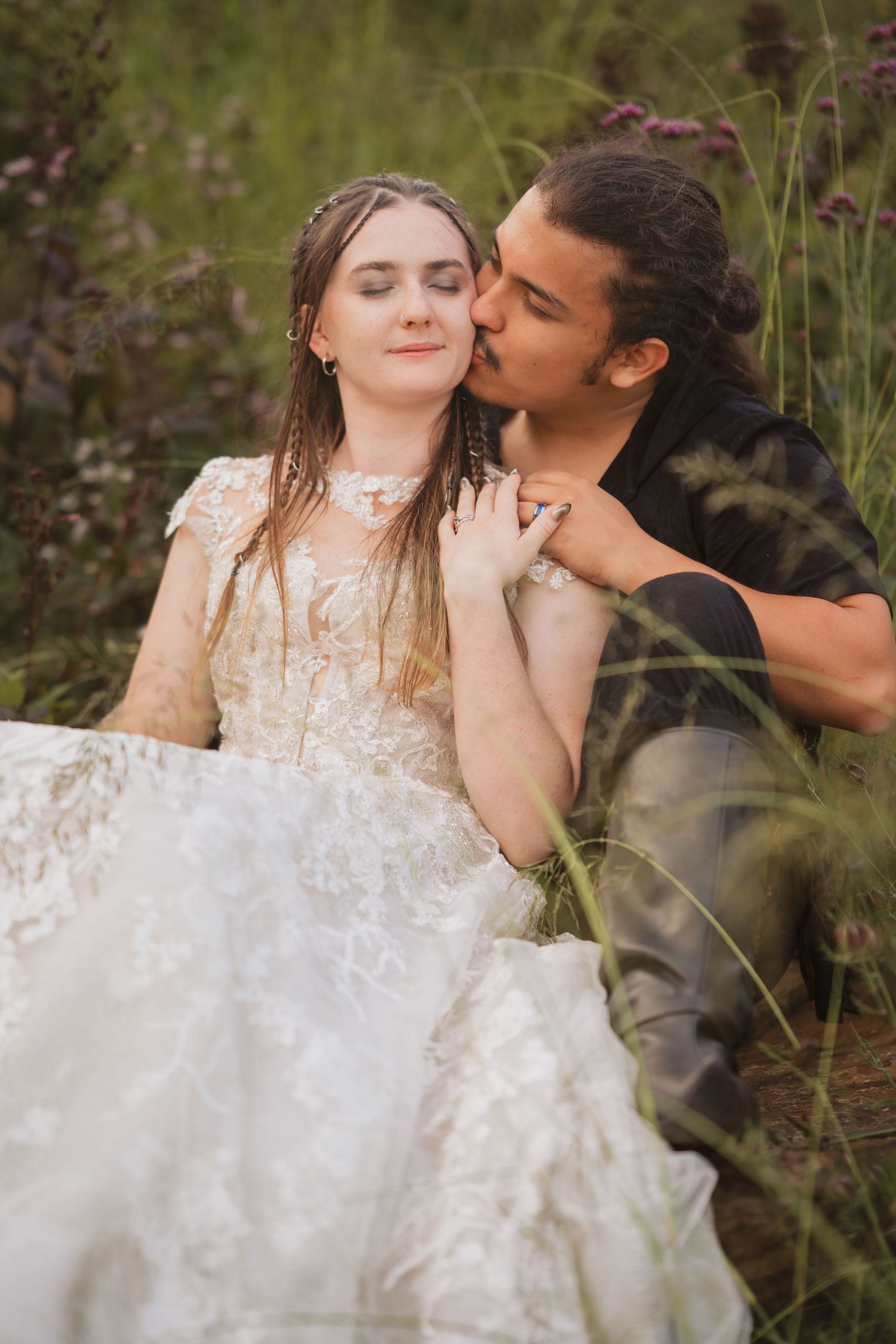 A romantic couple poses intimately in tall grass, with the bride wearing a detailed lace wedding gown.