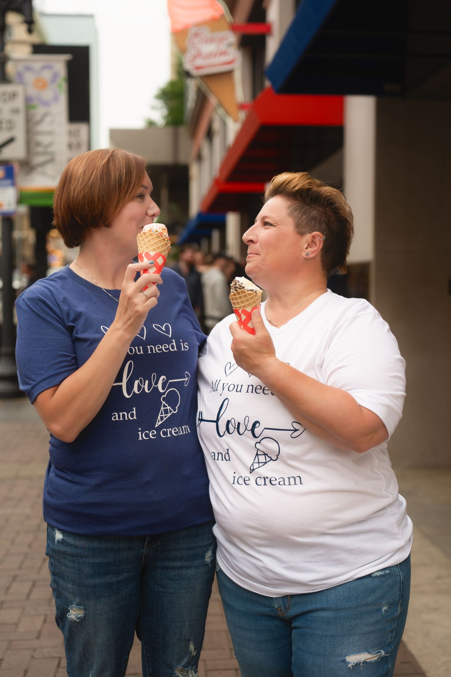 Two people in matching 'live love ice cream' t-shirts playfully share a sweet treat together on a city street.