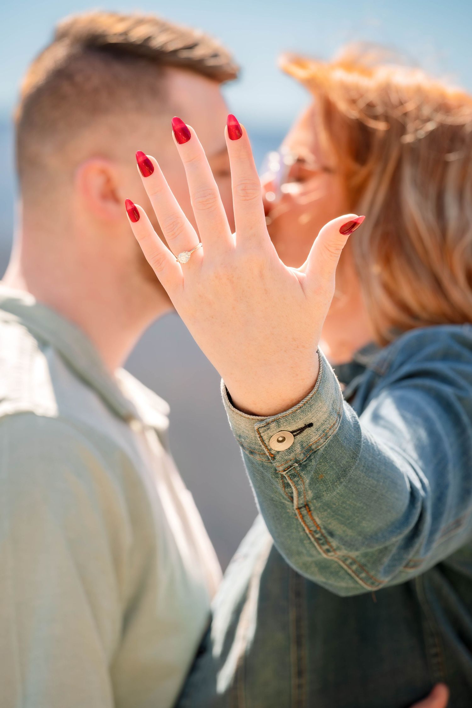 Woman in denim jacket waves hand with red nail polish against light blue background.
