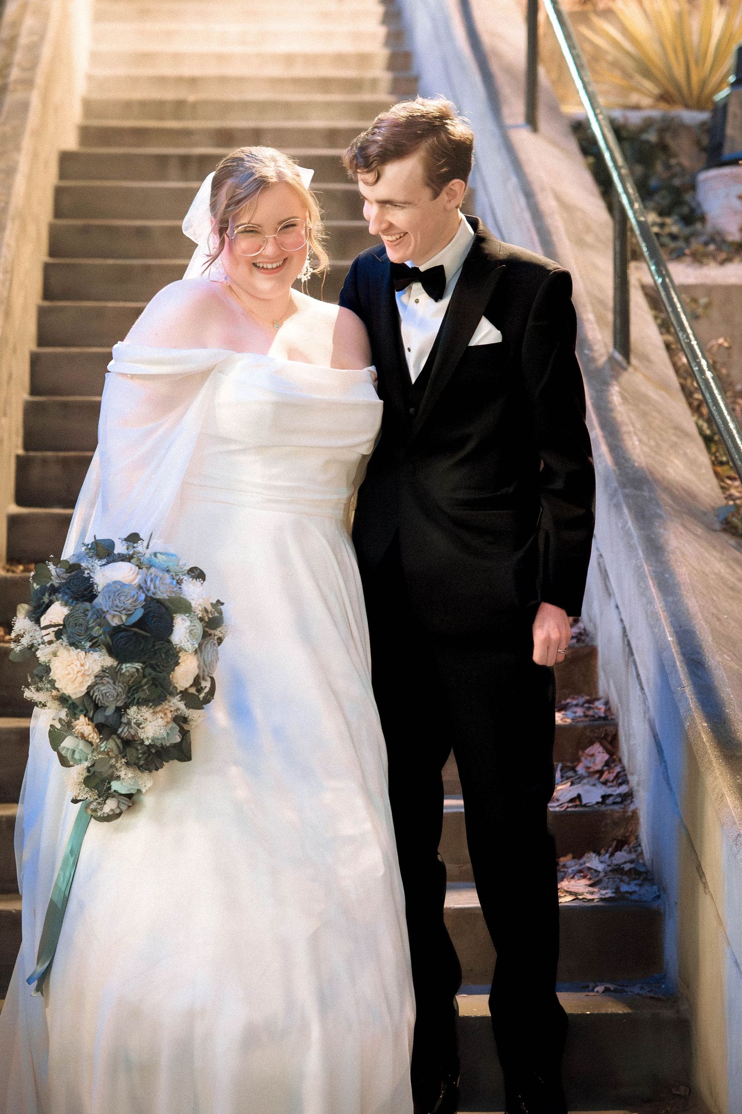 Happy couple share a joyful moment on stairs at their wedding, bride in white gown holding blue bouquet, groom in tuxedo.