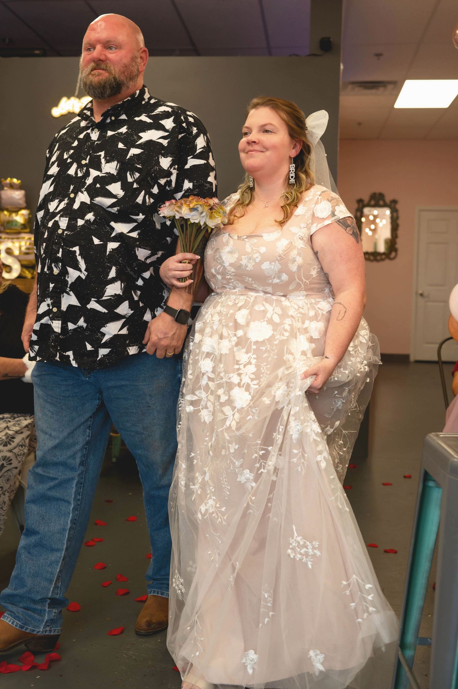 A wedding couple stands together at their reception, with the bride wearing a floral lace dress.