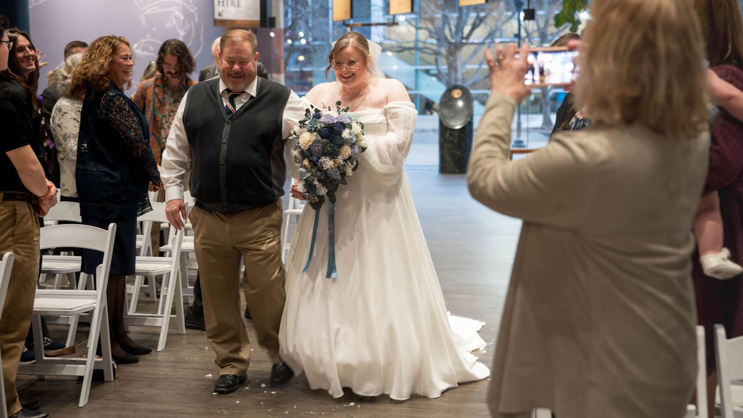 A wedding ceremony takes place in an indoor venue with guests seated in white chairs watching someone in a flowing white gown.