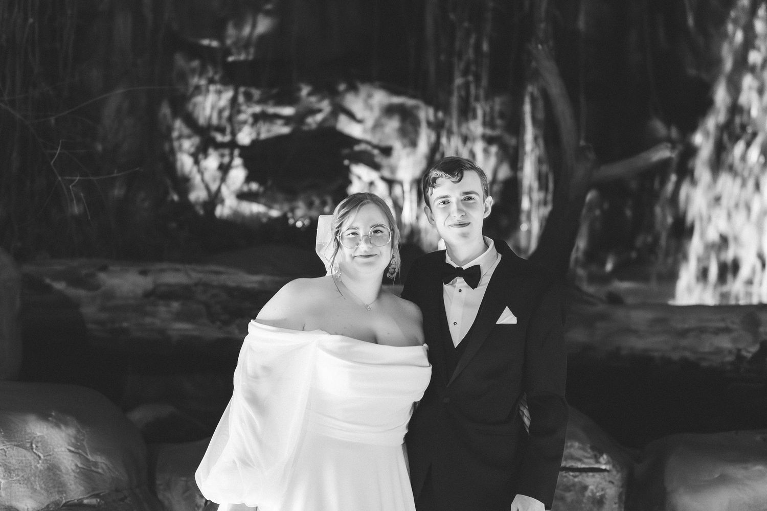 Black and white wedding photo of a couple outdoors, with the bride wearing an off-shoulder dress and groom in a tuxedo.