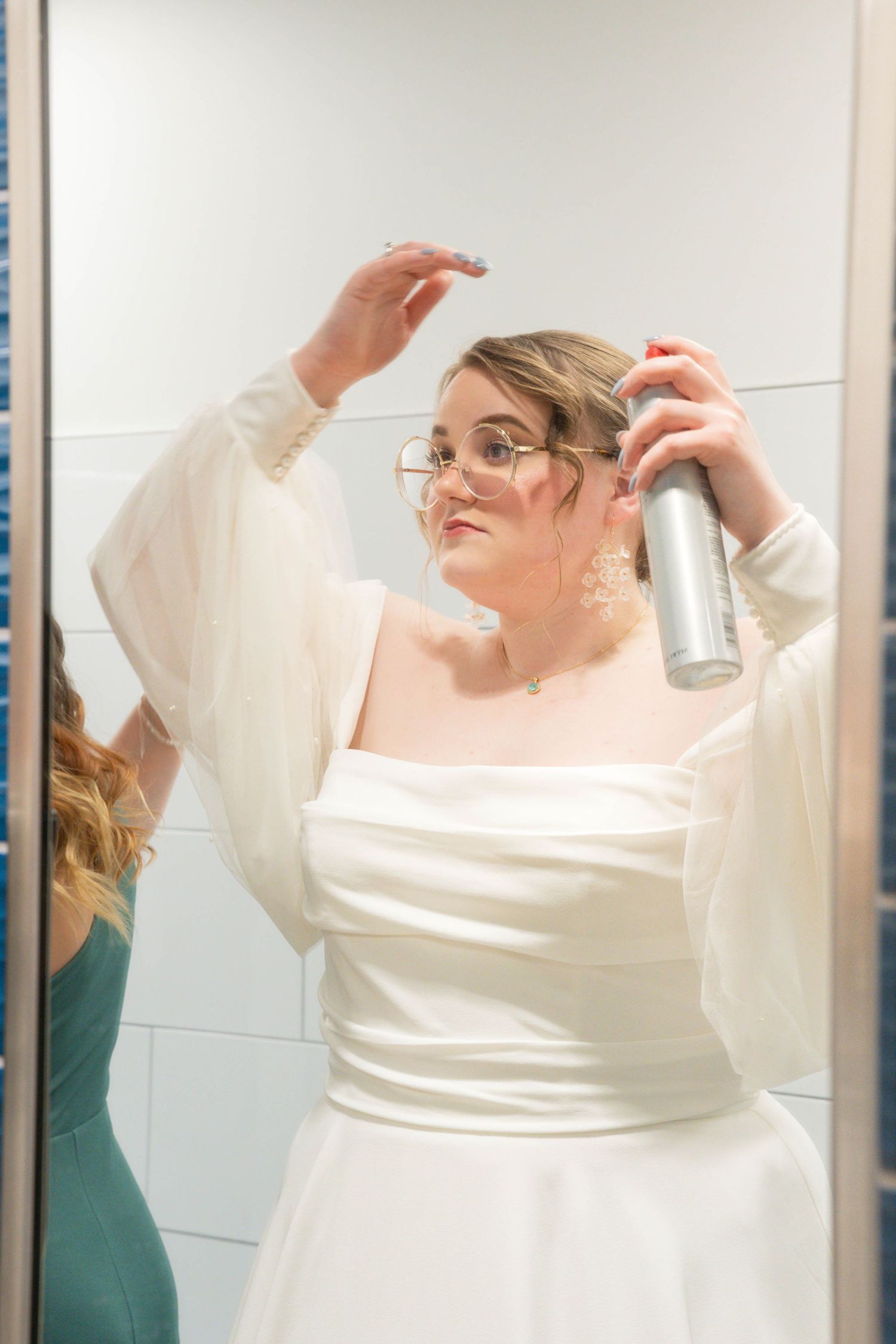 Someone in a white wedding dress with billowing sleeves applies hairspray while getting ready in a bathroom mirror.