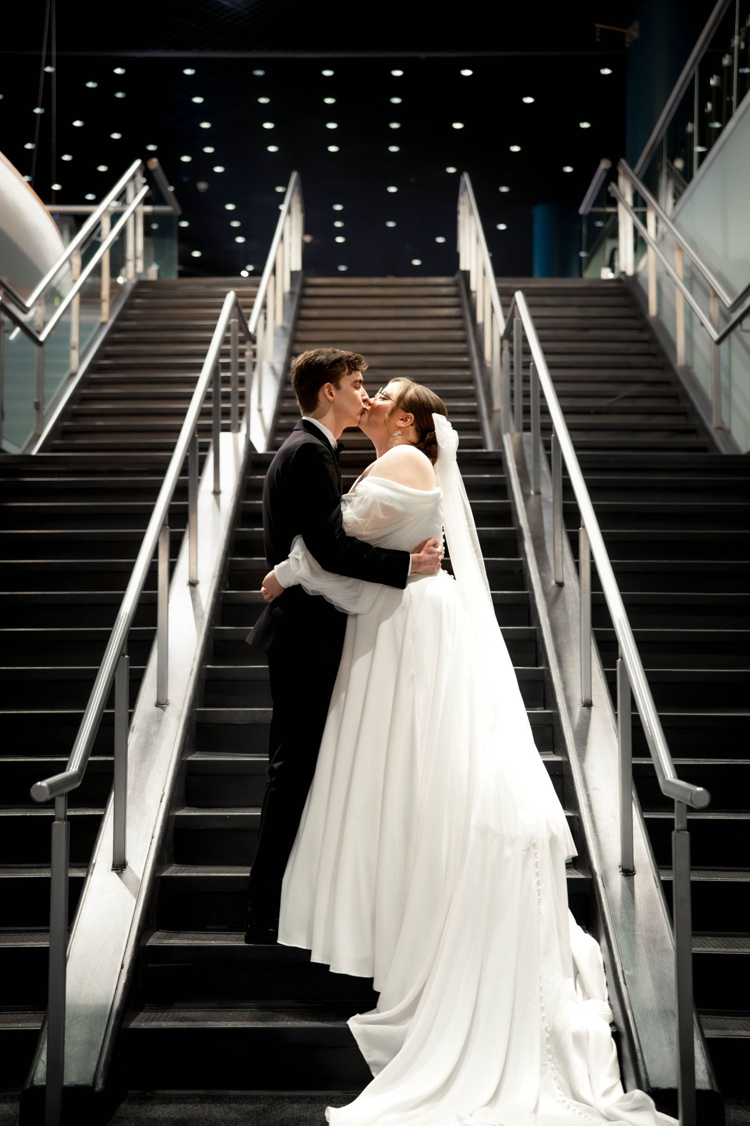 A couple in wedding attire share a romantic kiss on a grand modern staircase with dramatic lighting and white handrails.