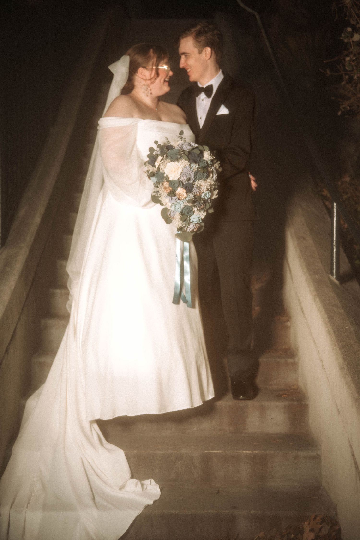 A vintage-style wedding portrait of a couple standing on stairs with the bride holding a floral bouquet.