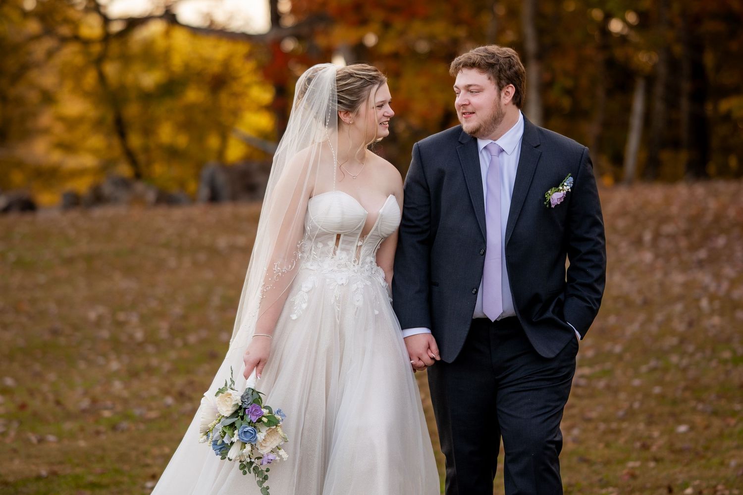 A couple in wedding attire shares a romantic moment among vibrant autumn foliage in a forest setting.