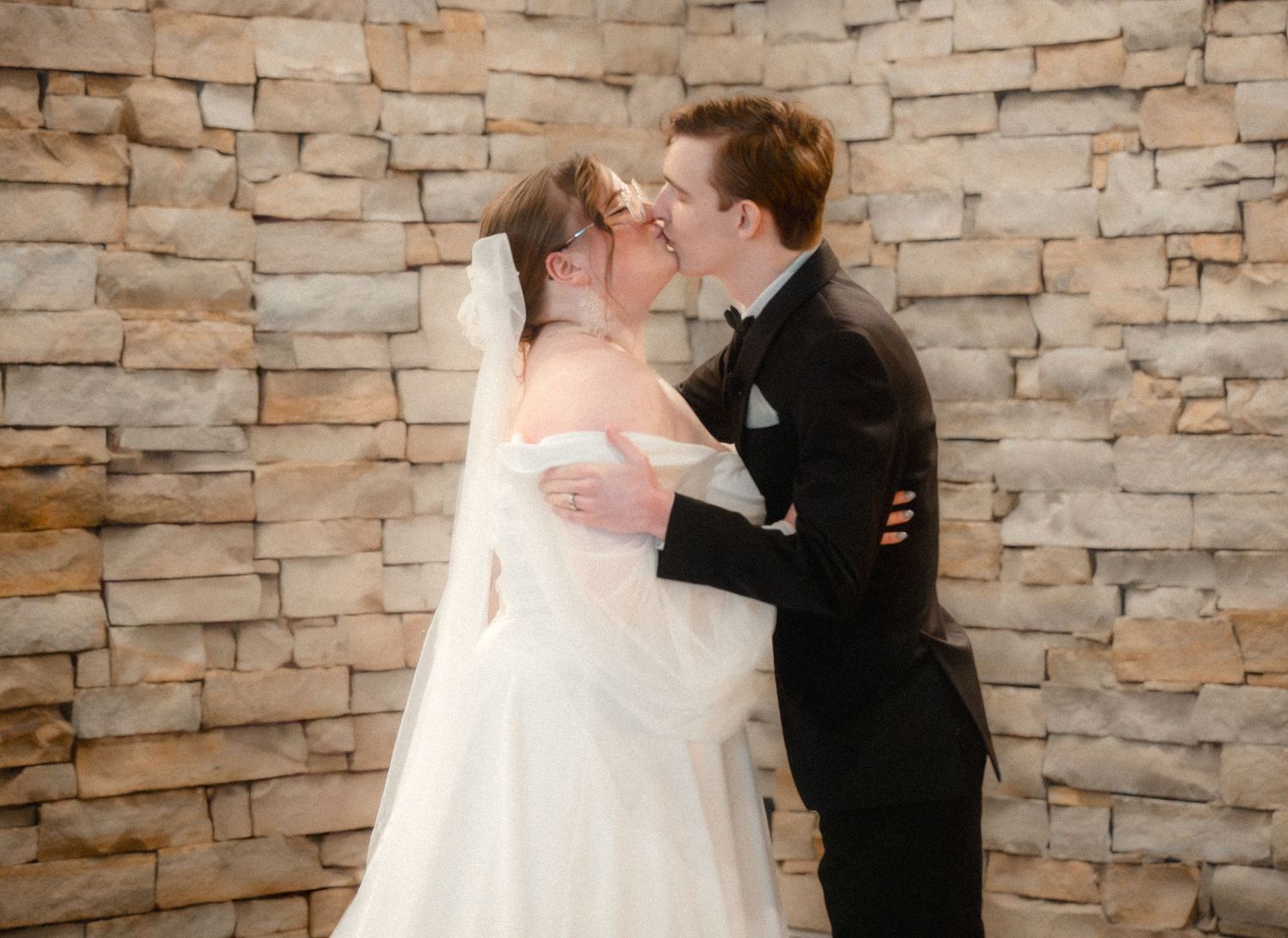 A romantic wedding moment between a couple in formal attire embracing against a stone wall backdrop during their ceremony.