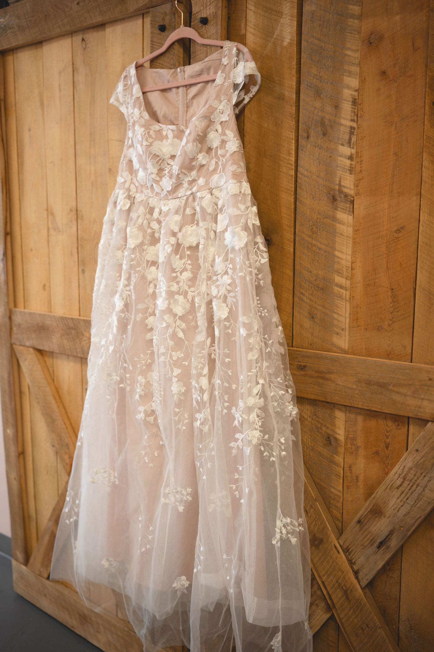 A delicate blush wedding dress with floral lace appliques hangs on a wooden hanger against rustic barn walls.
