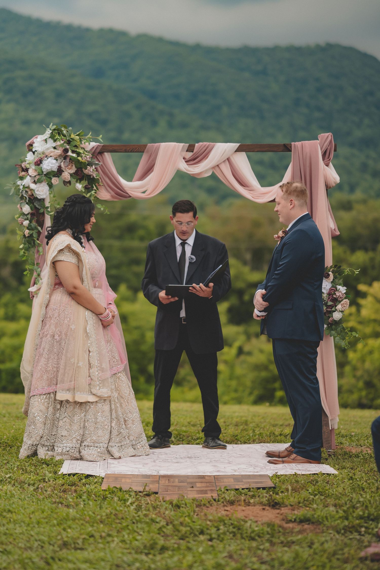 An outdoor fusion wedding ceremony with pink and white decor against a scenic mountain backdrop.