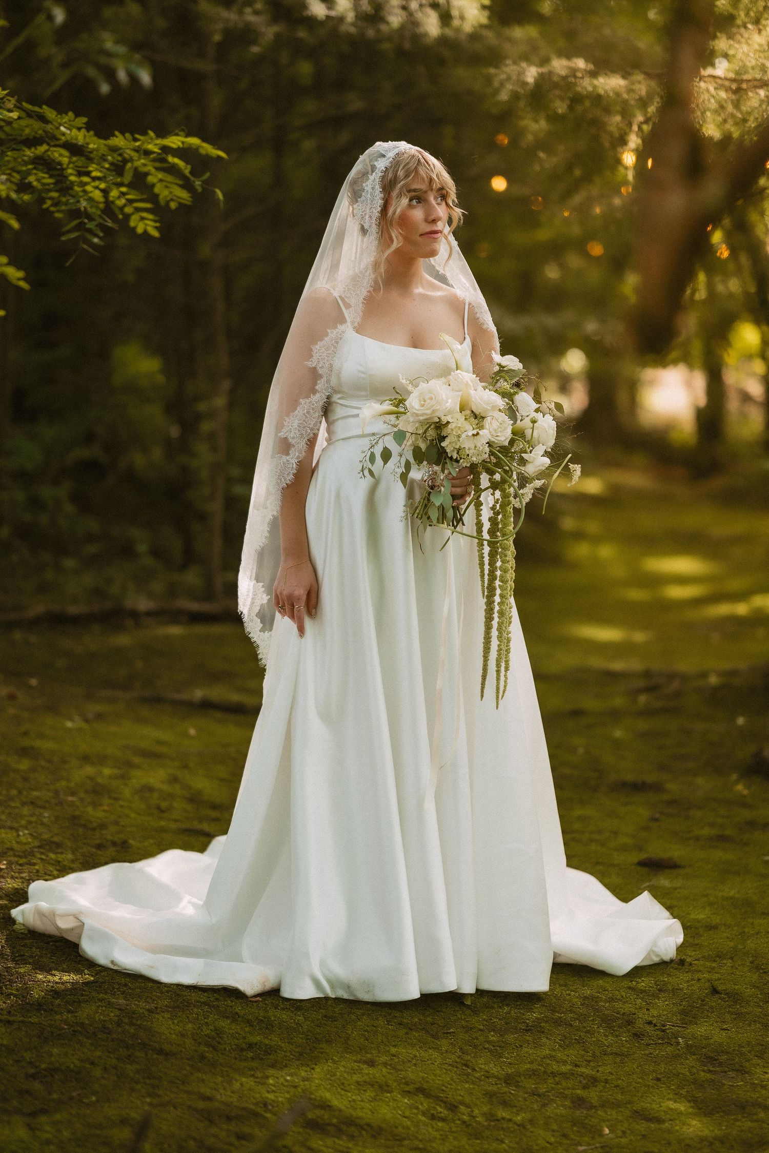 An elegant bridal portrait features a white wedding dress against lush green woodland scenery.