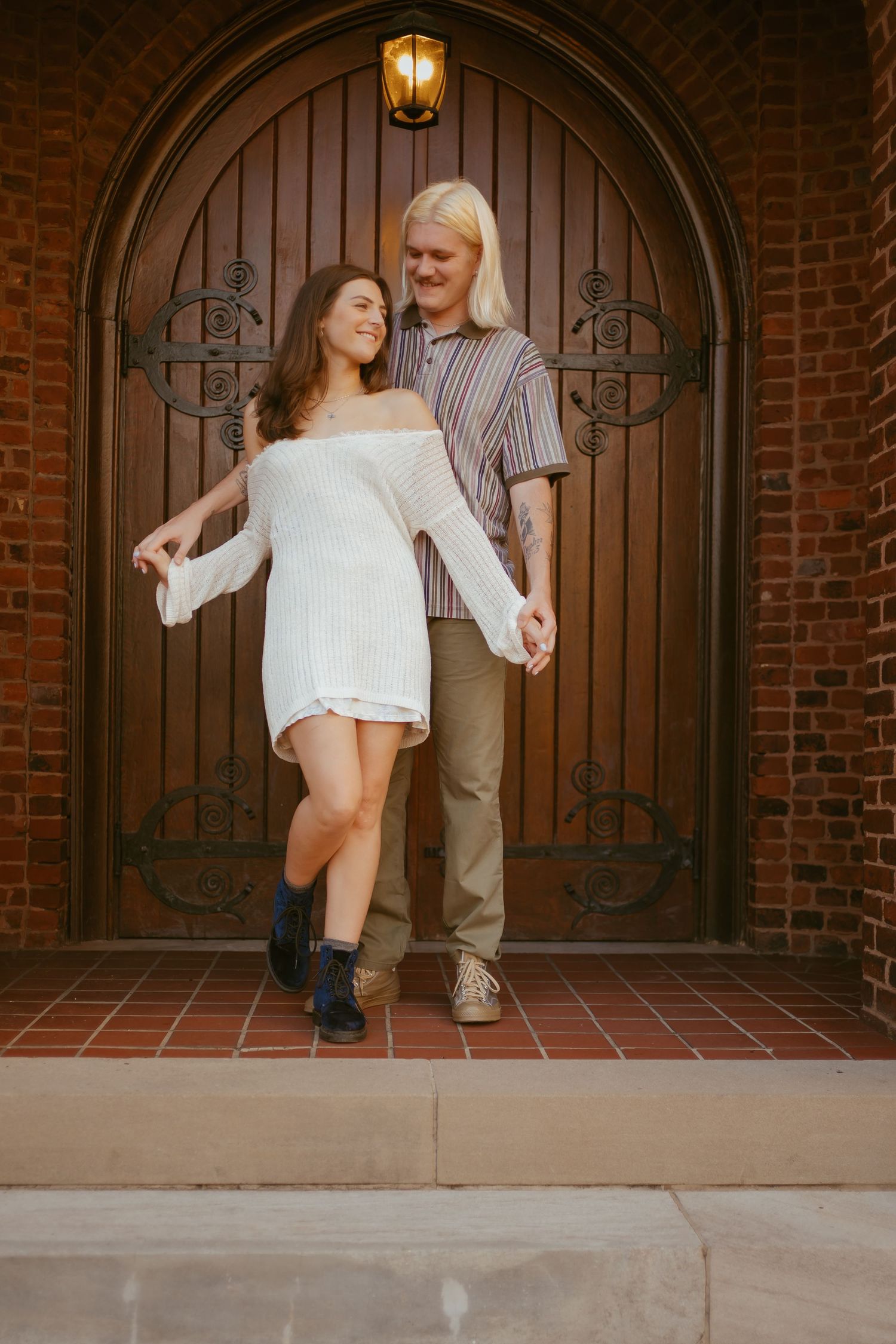 A romantic couple enjoys a playful dance session in front of an arched wooden door against a brick wall.