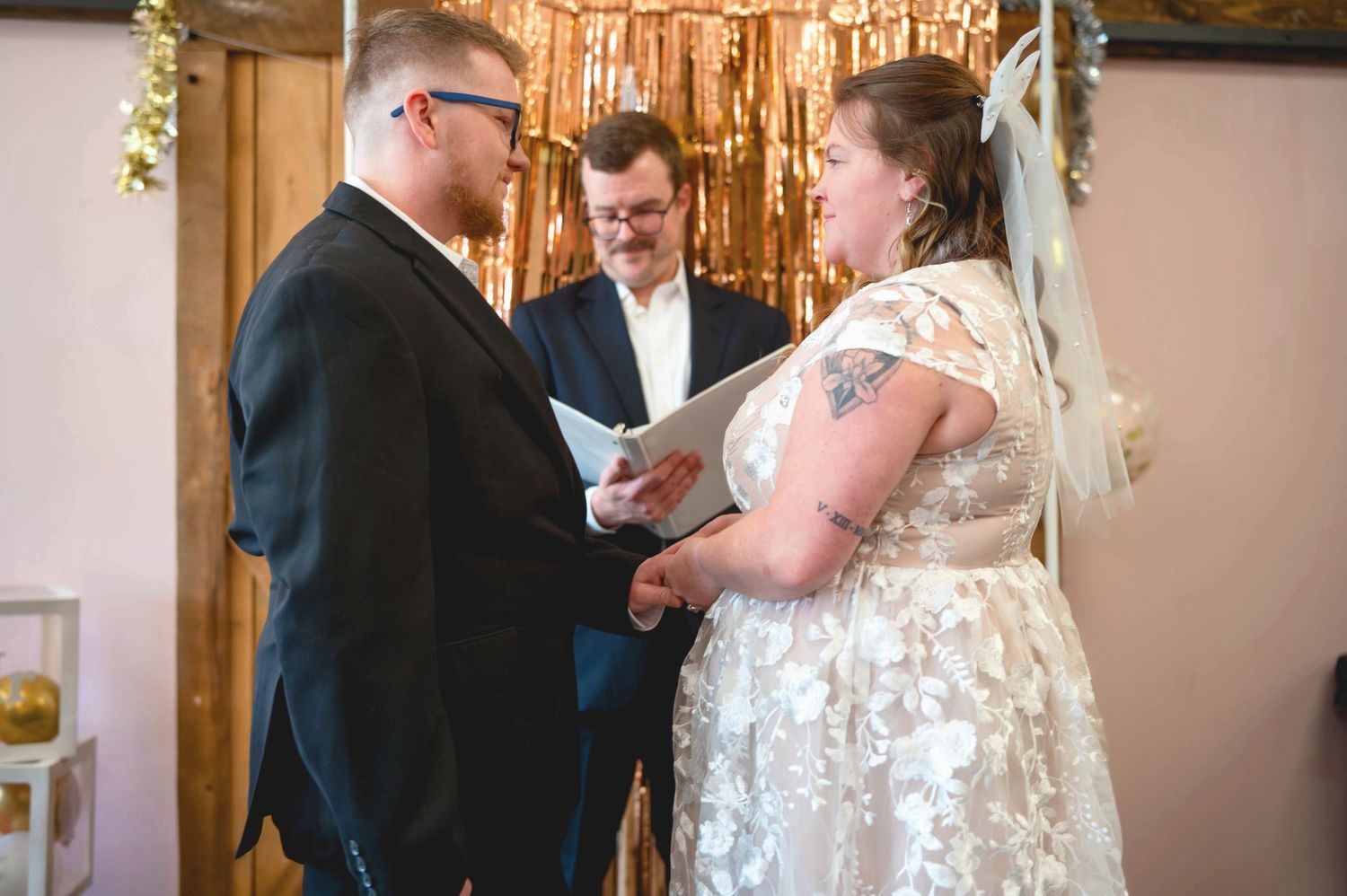 A wedding ceremony takes place indoors with a couple exchanging vows in front of an officiant beneath decorative lights.