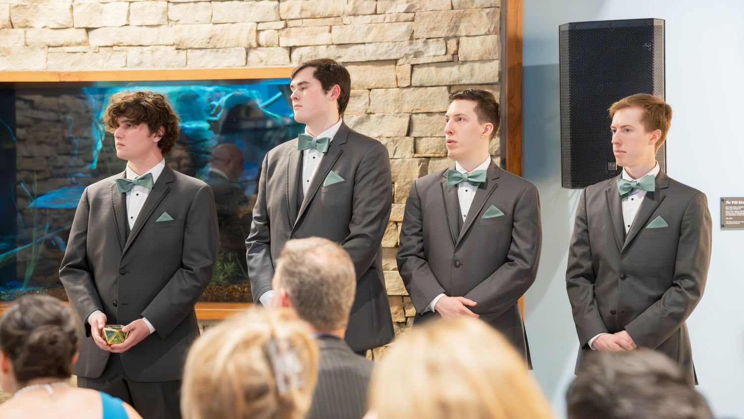 The groomsmen in charcoal gray suits stand in a row at the ceremony, waiting attentively near a stone wall.
