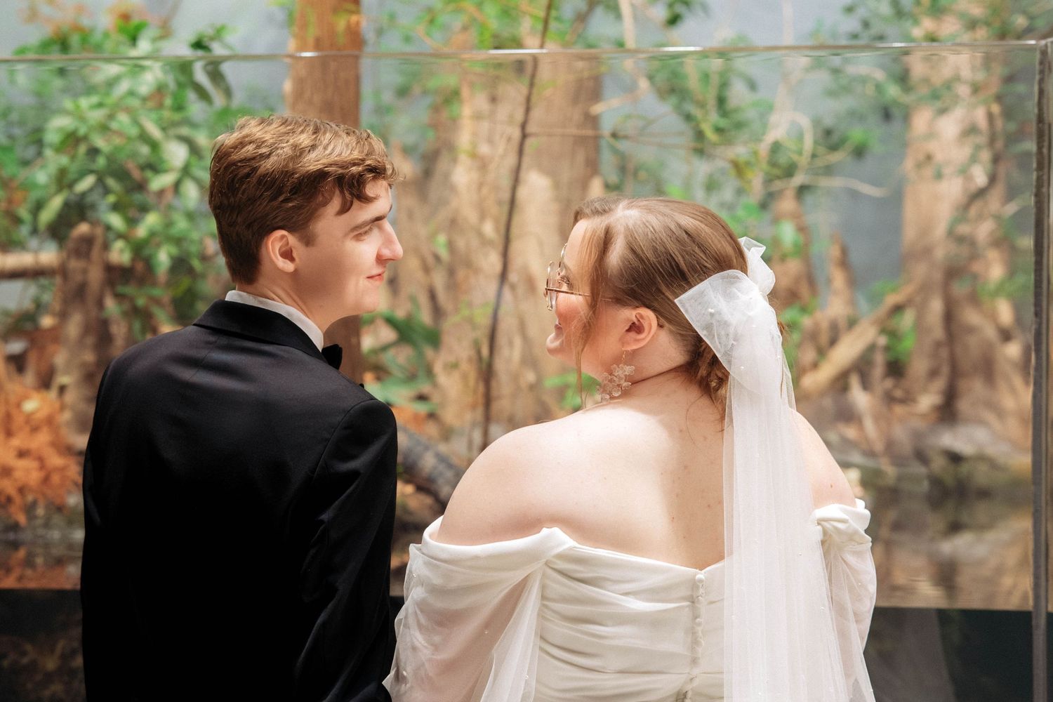 Bride in off-shoulder dress and groom in black suit share romantic moment in natural setting.