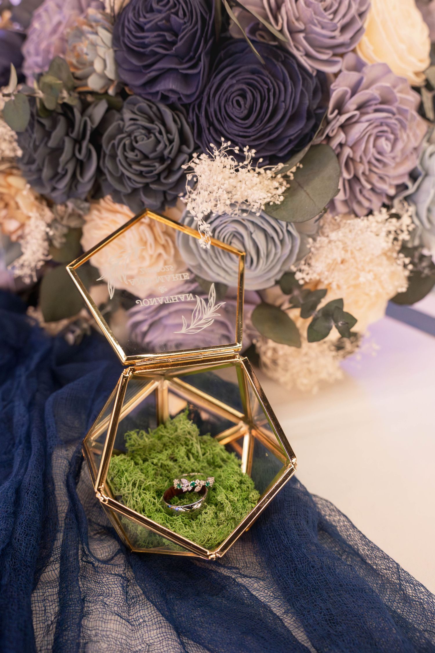 Wedding rings displayed in gold geometric terrarium with moss, alongside purple and mauve roses and baby's breath bouquet.